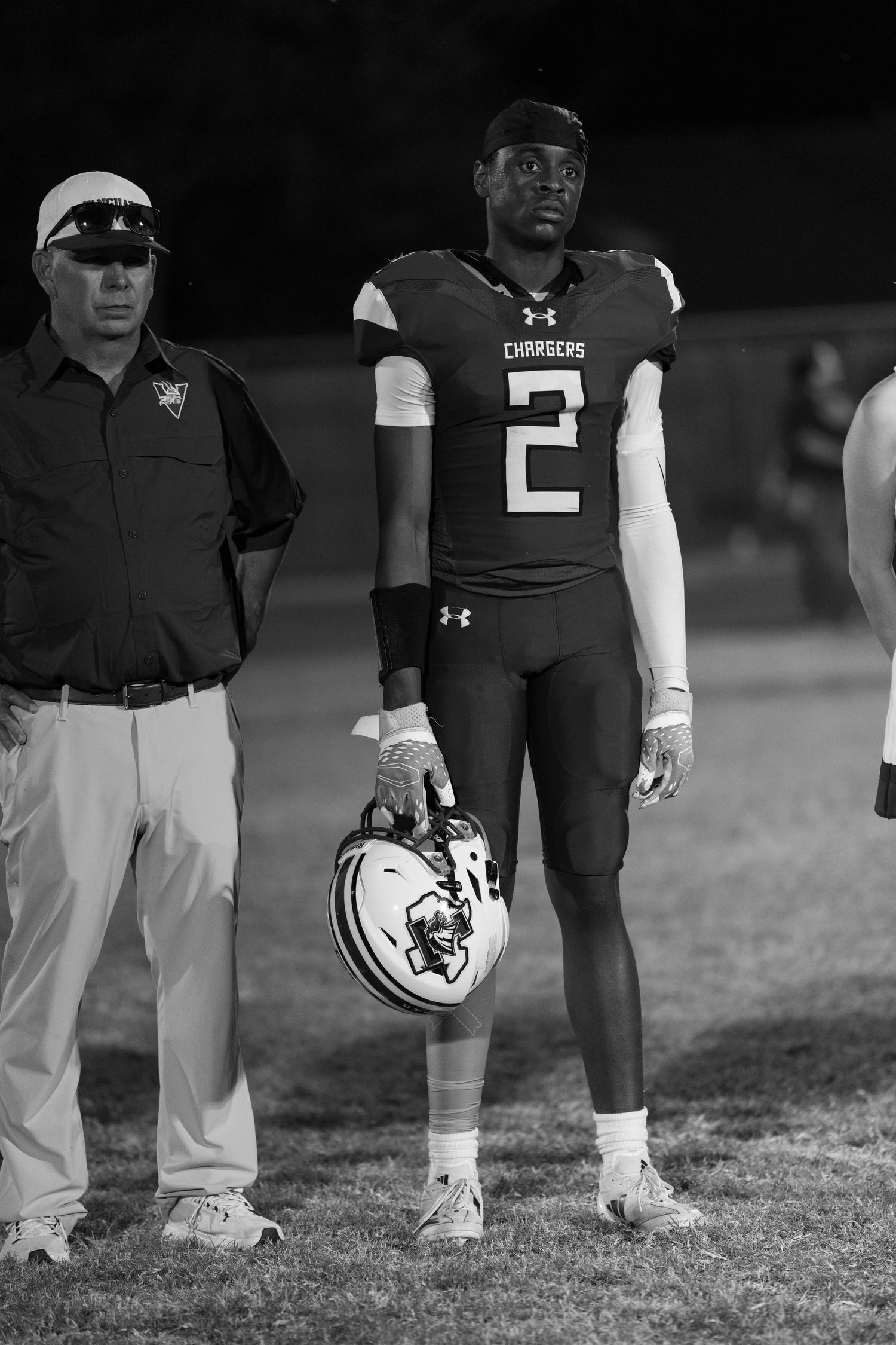 Football player in black uniform holds helmet while standing on field during nighttime game.