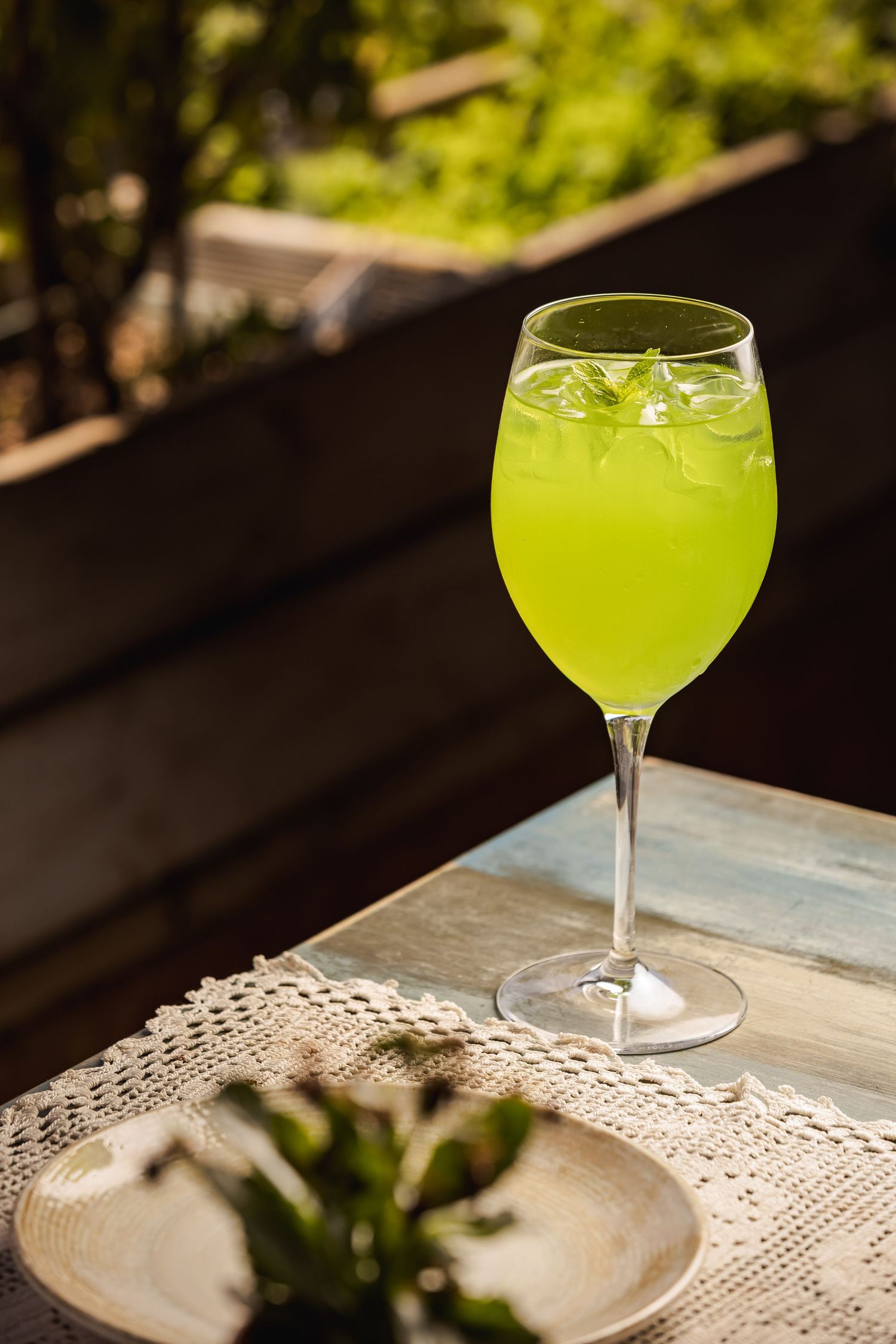 A bright yellow-green cocktail served in an elegant coupe glass sits on a white lace tablecloth in natural lighting.
