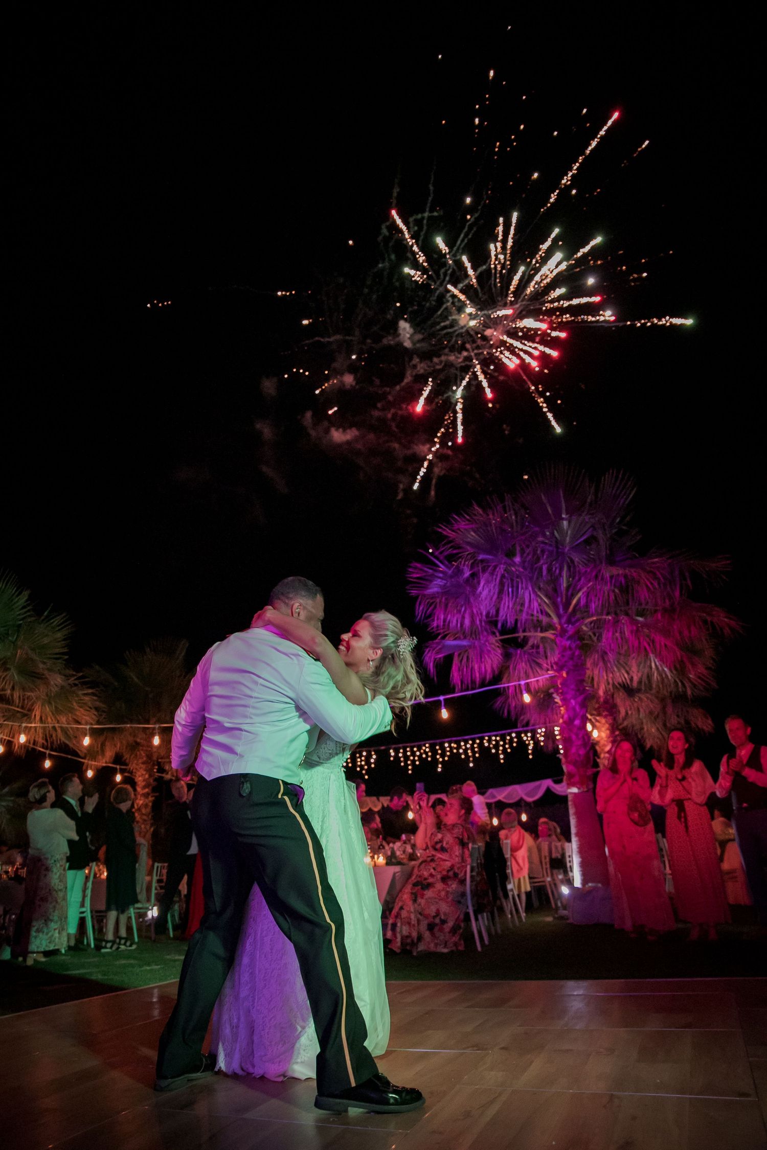 Newlyweds dancing beneath colorful fireworks and string lights at outdoor evening reception.