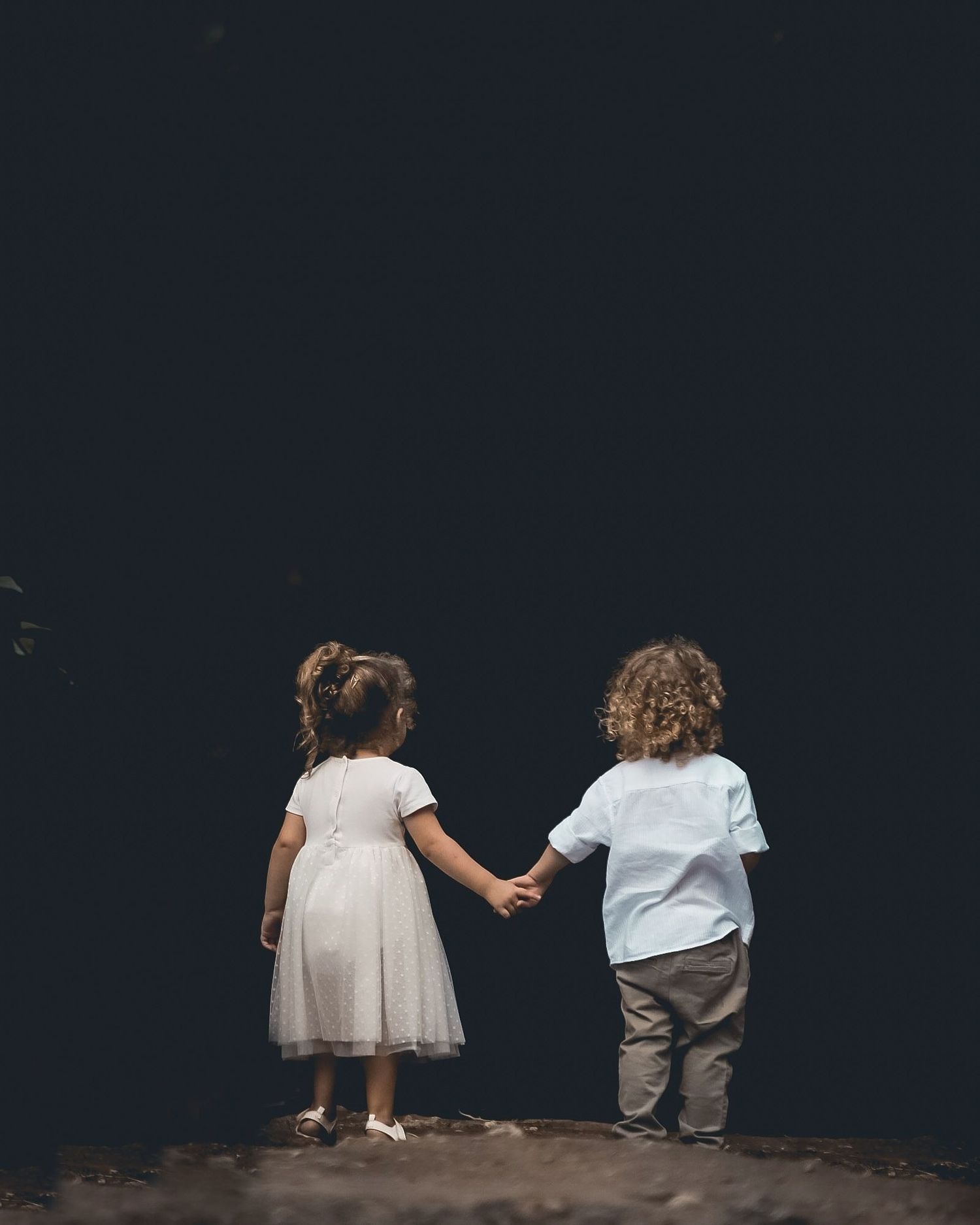 Two small children sit side by side in white and gray outfits against a dark background.
