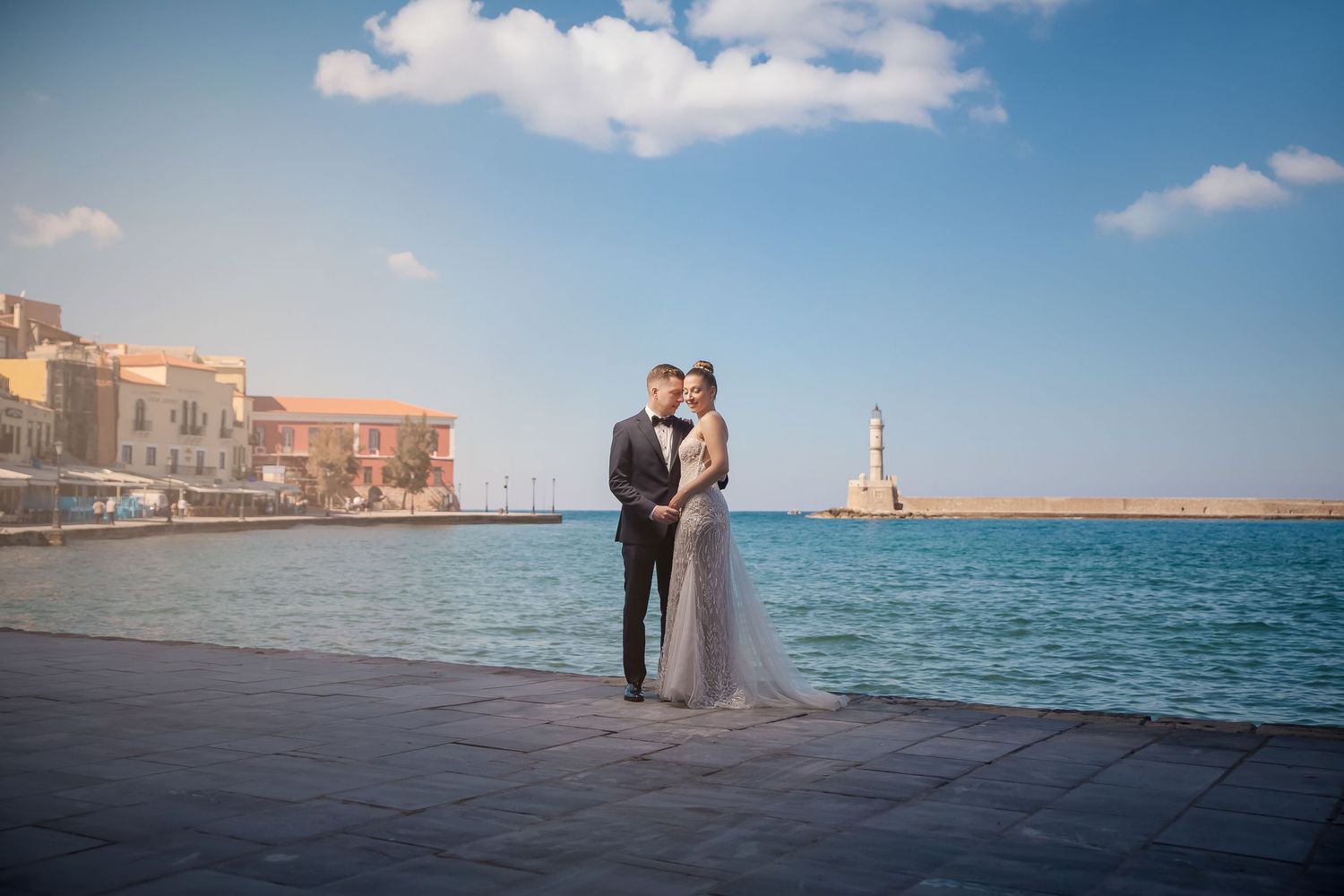 Wedding couple posing by Mediterranean waterfront with historic architecture and lighthouse in the background.