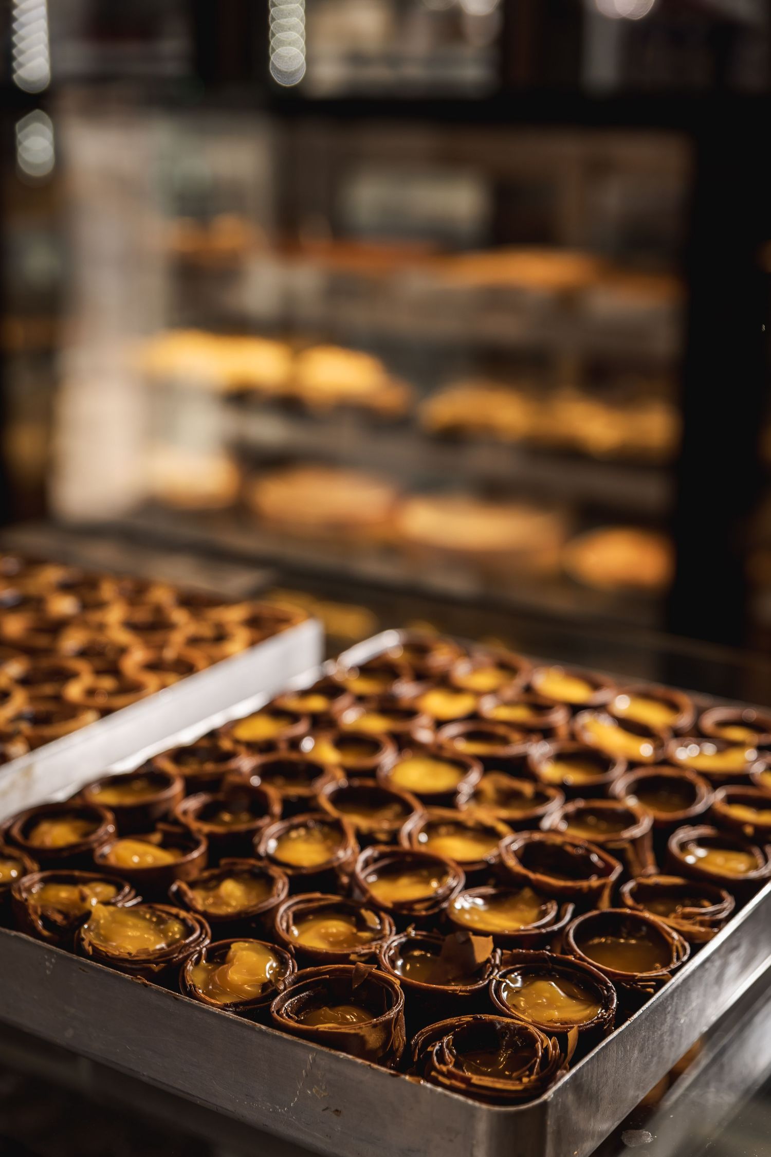 Rows of freshly baked Portuguese custard tarts with caramelized tops cooling on metal baking trays in a bakery display case.
