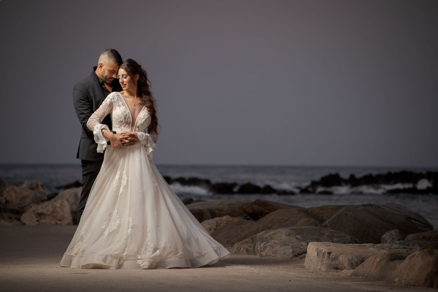 A couple wearing wedding attire poses for romantic sunset photos on a rocky beach.