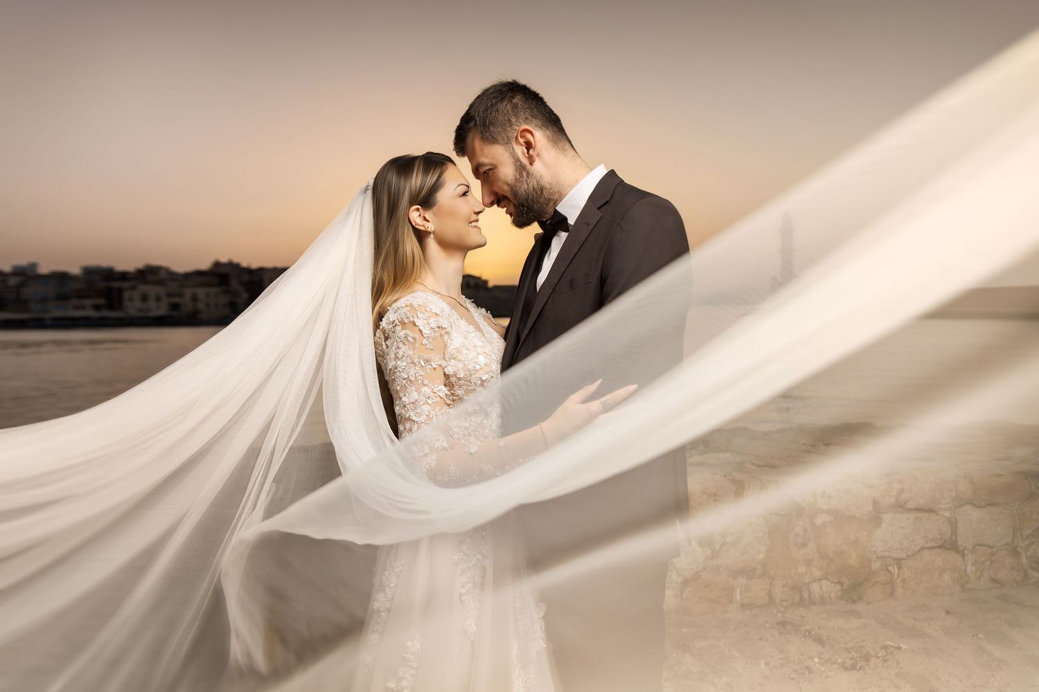 Elegant wedding couple poses under flowing white veil against desert background at sunset.