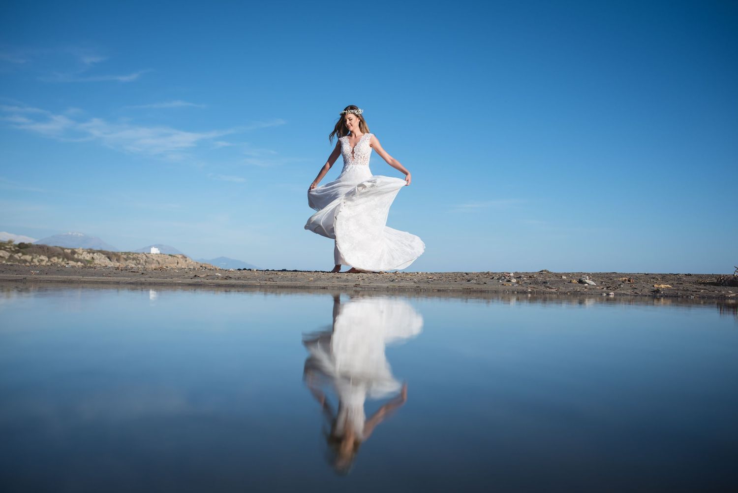 Bride in flowing white dress walks along serene lakeside during sunset photo session.