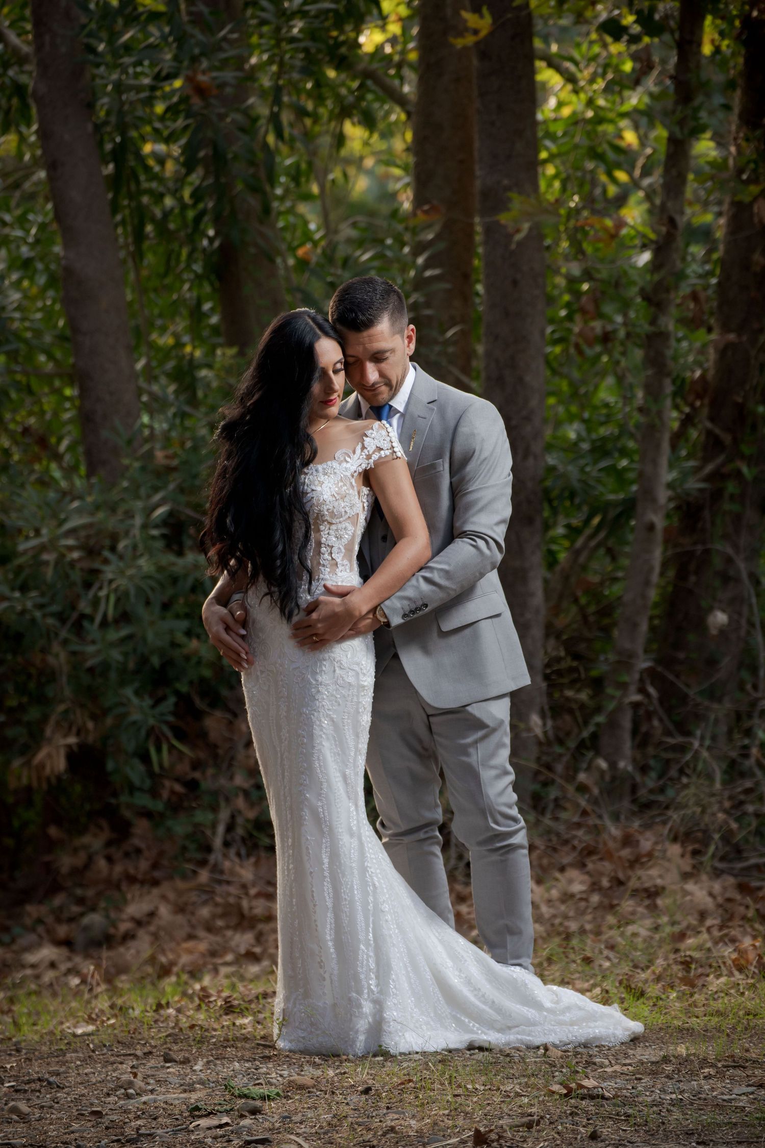 Wedding couple embraces in romantic woodland setting wearing traditional white dress and grey suit.