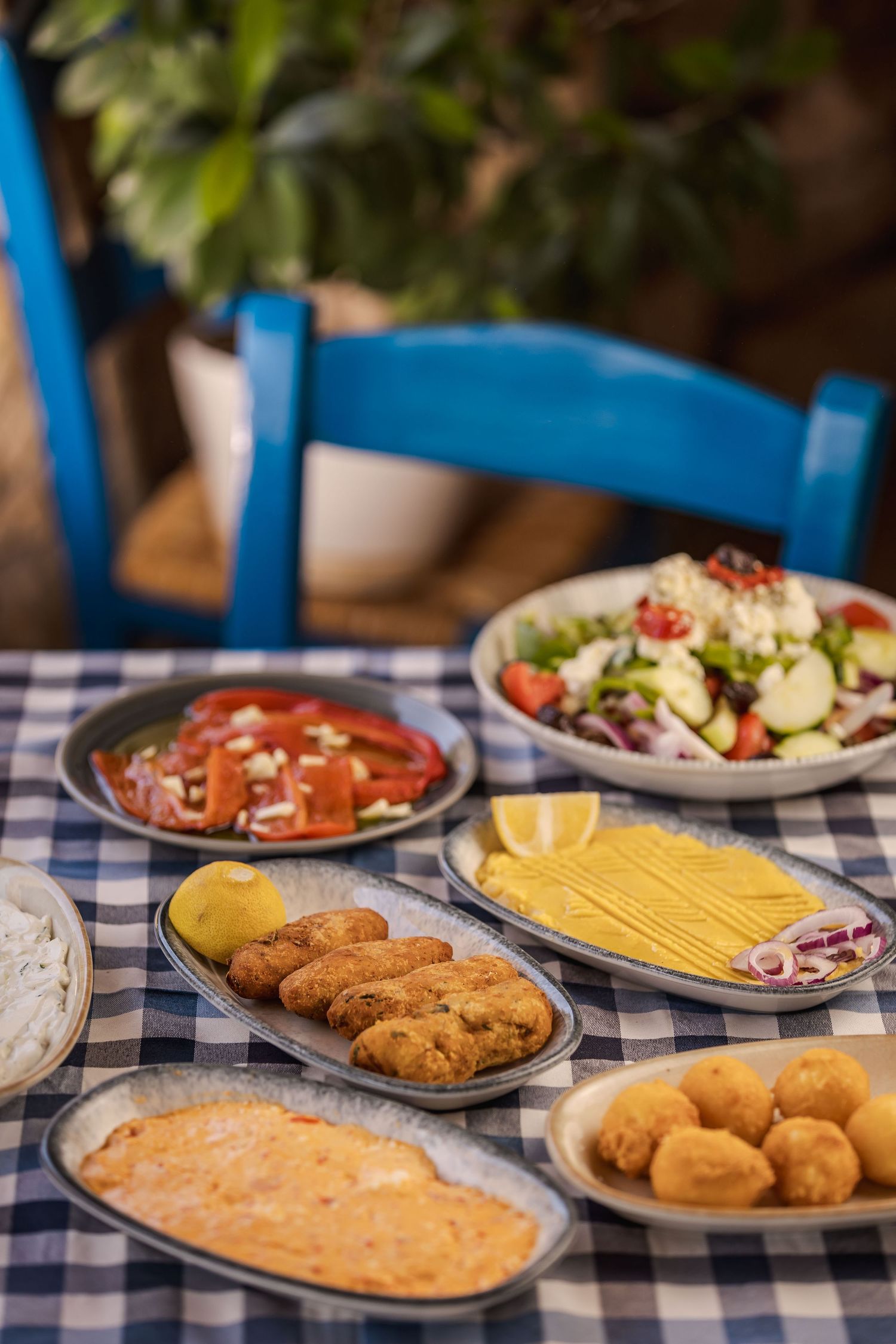 Mediterranean appetizer spread with fried croquettes, pasta, fresh salad, and dipping sauces on a blue checkered tablecloth.