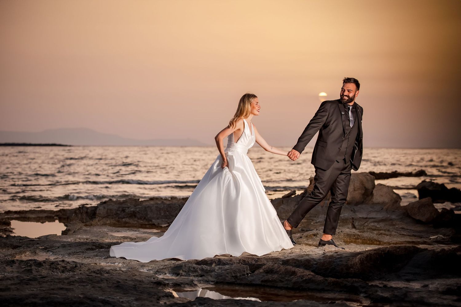 Romantic seaside wedding photos at sunset with couple walking along rocky coastline.