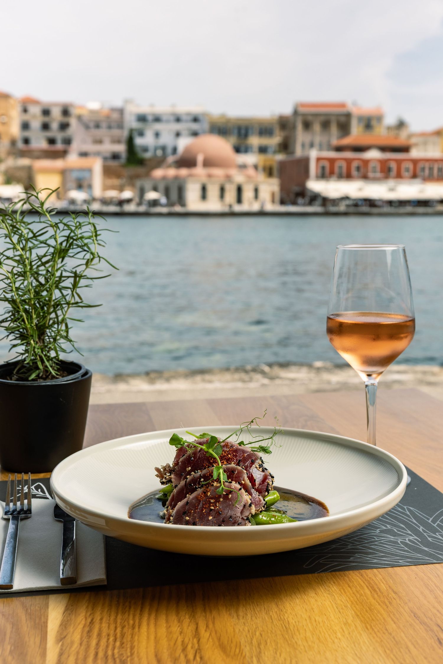 Elegant dinner plate with meat dish beside rosé wine glass overlooking colorful Mediterranean waterfront buildings.