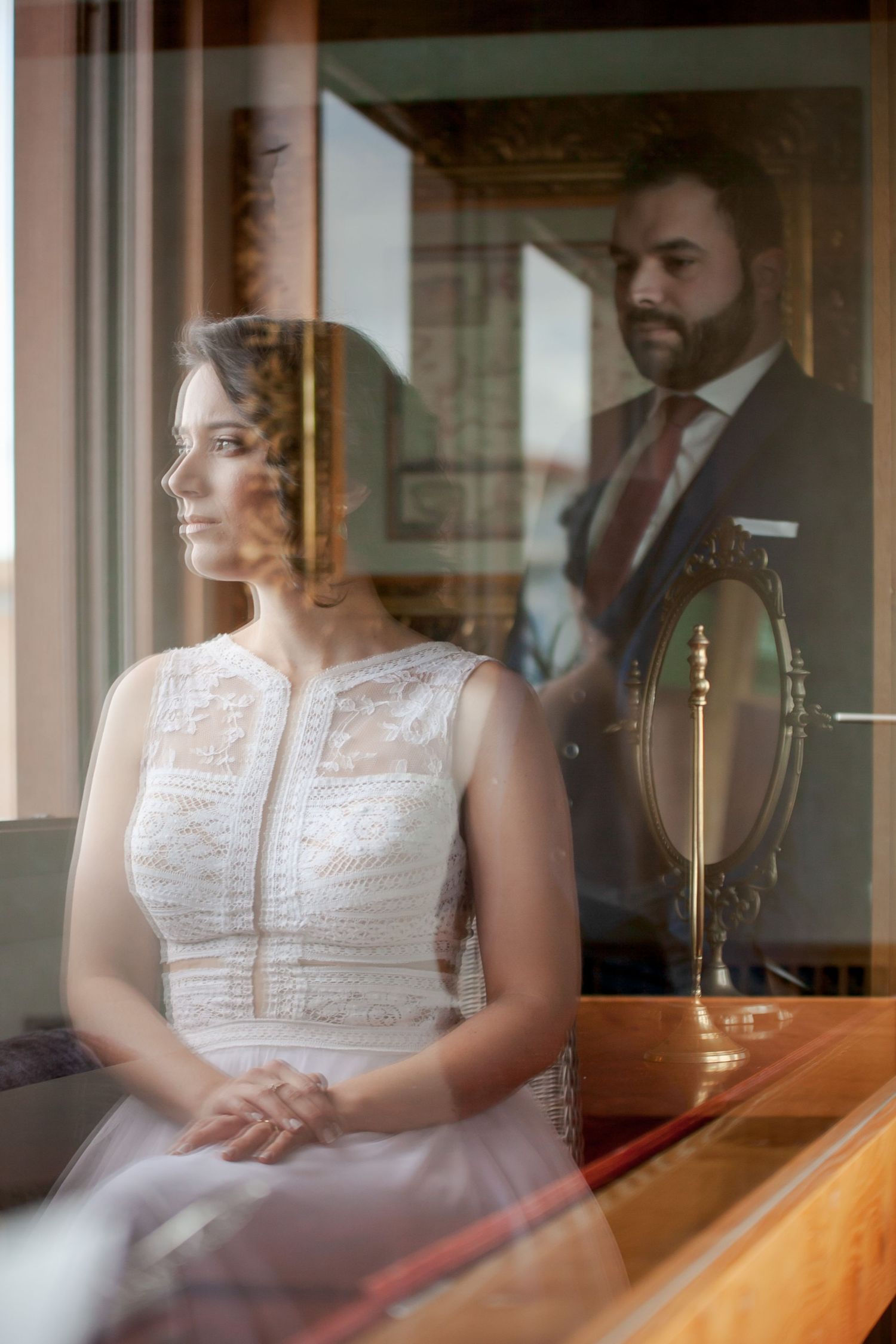Artistic reflection through glass shows bride in white dress seated in elegant indoor setting.