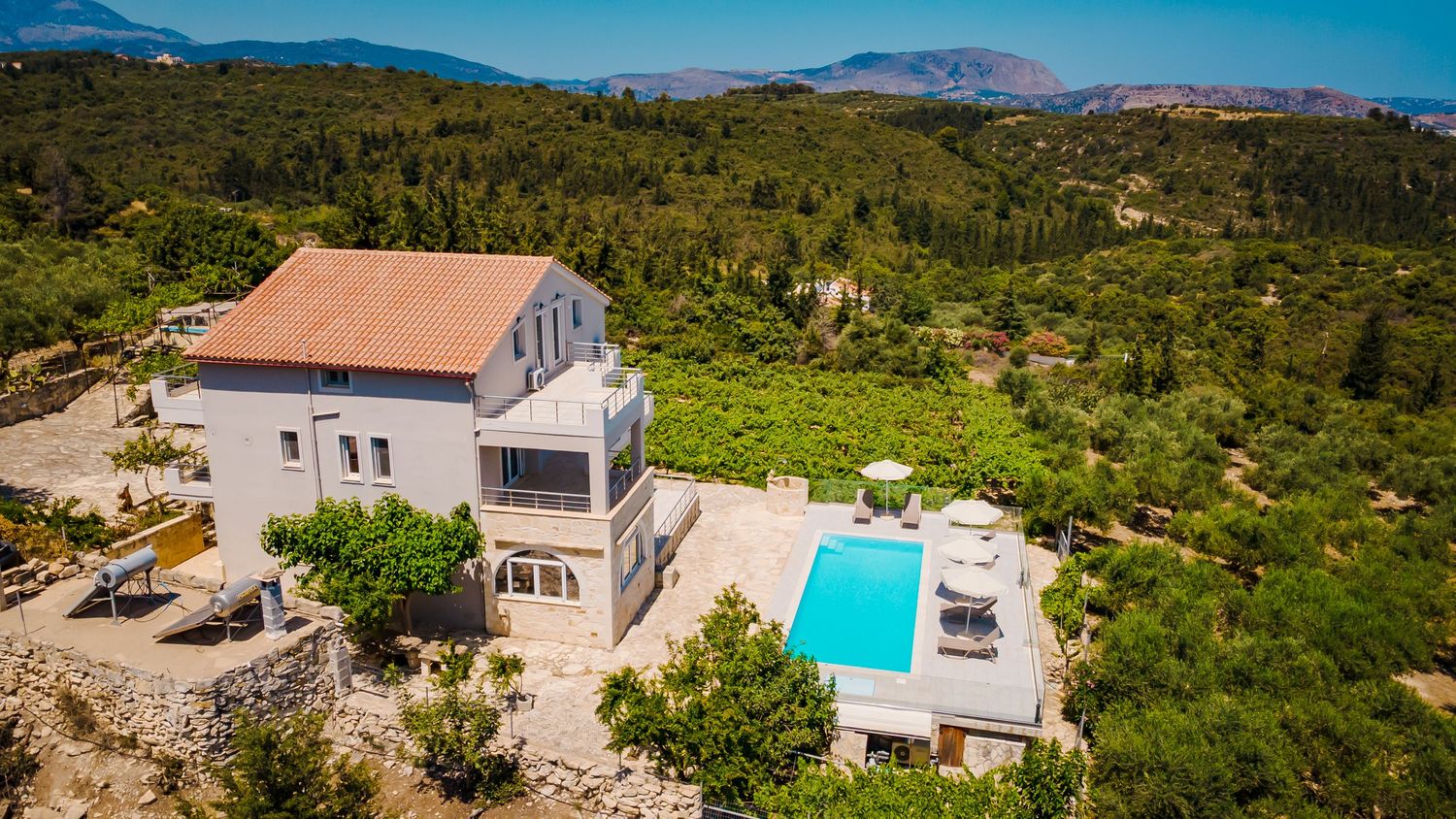 Scenic aerial panorama of coastal property with swimming pool and Mediterranean architecture nestled in lush hillside forest.