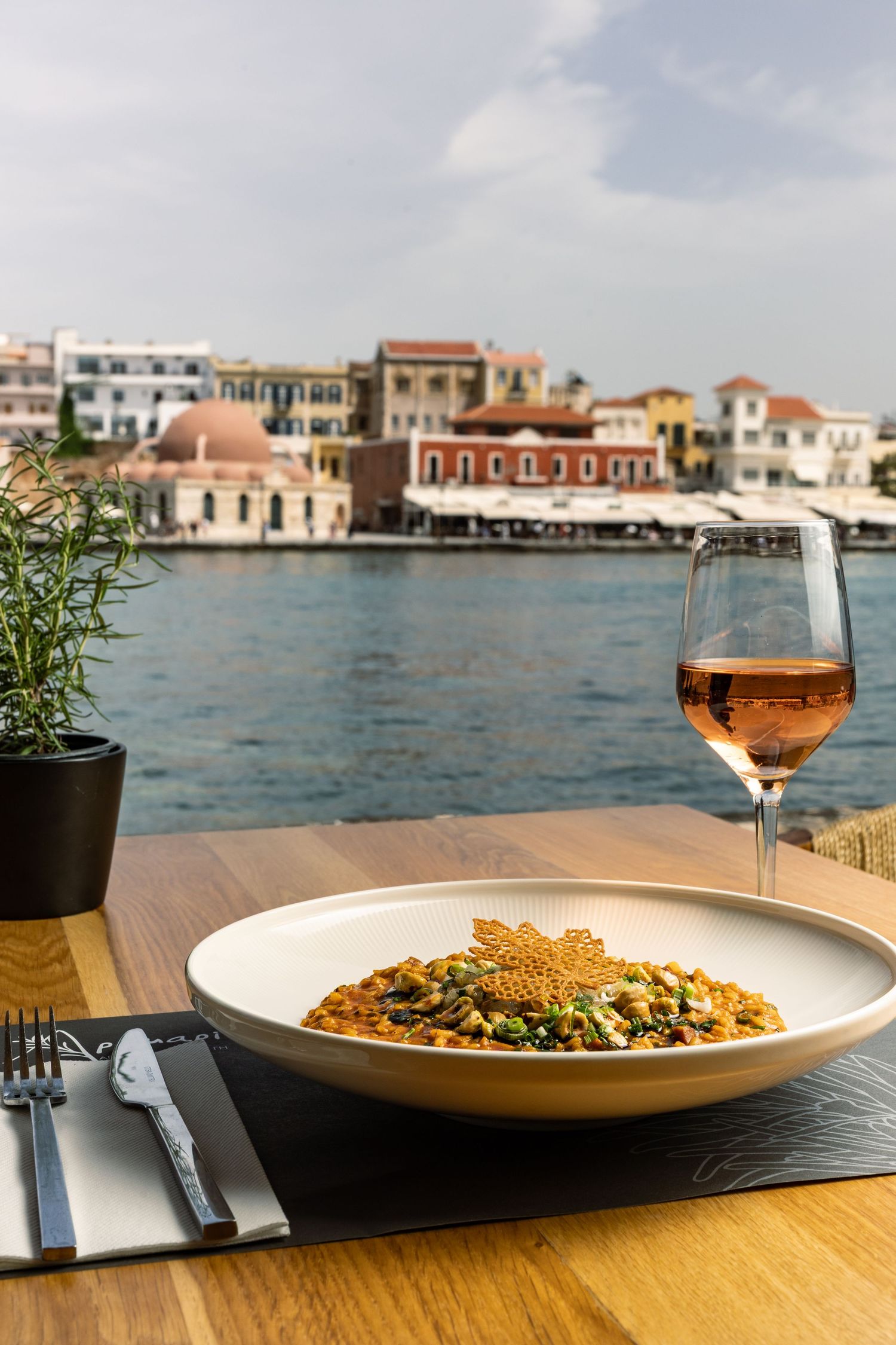 Elegant dining setup with pasta dishes and wine glasses overlooking a scenic waterfront with Mediterranean buildings.