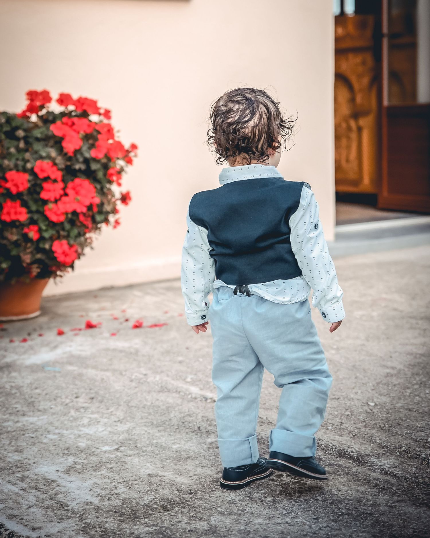 Small child in vintage-style outfit with ruffles walking on stone patio near red flowers.