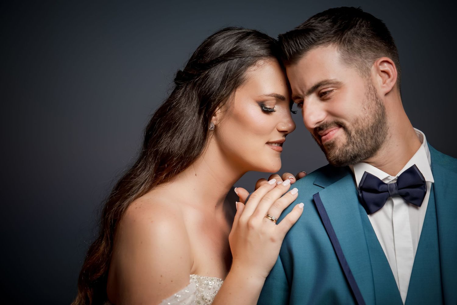Intimate close-up portrait of a couple in formal attire sharing a tender moment against dark background.