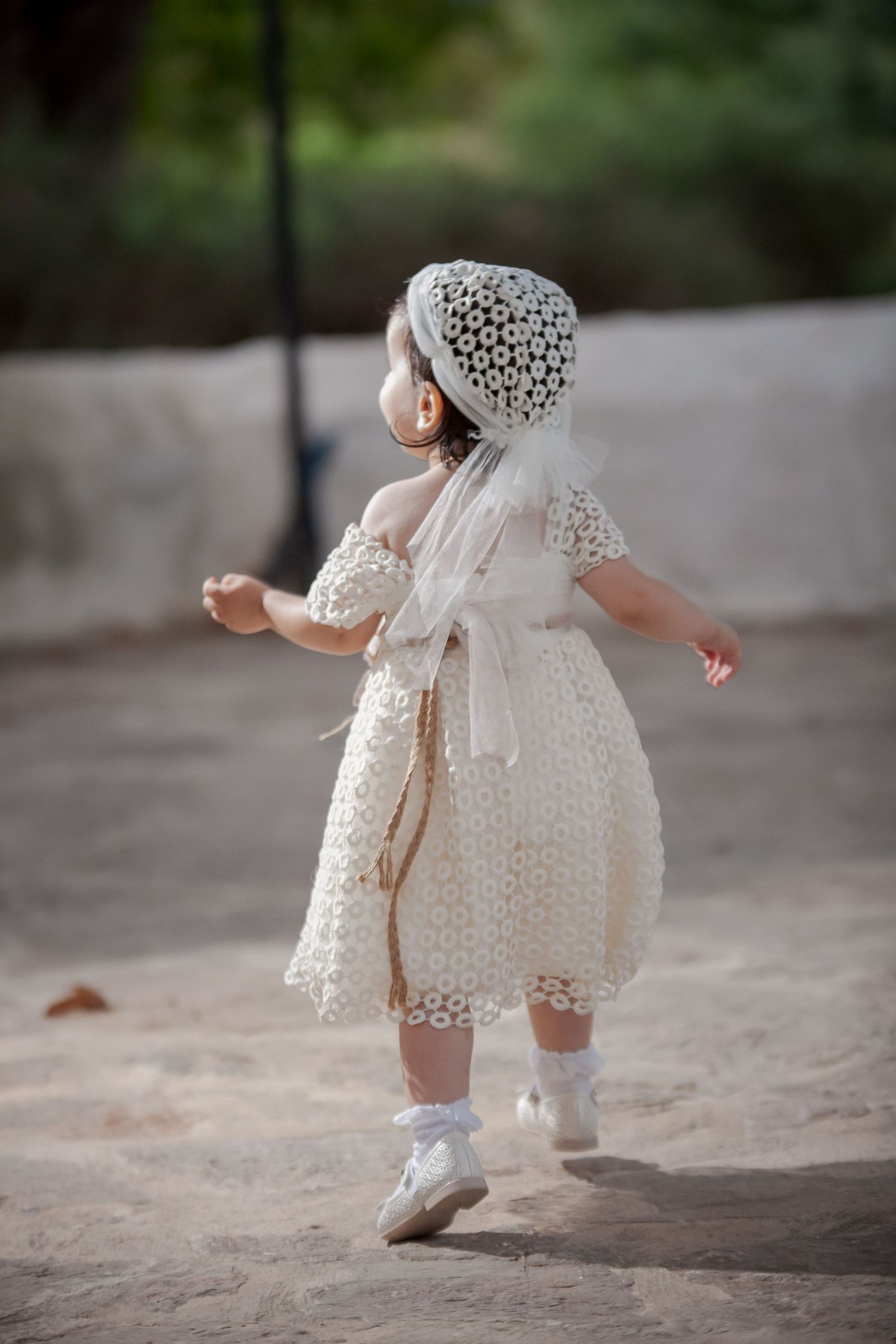 Small child in white eyelet lace dress and shoes walking on stone pavement.