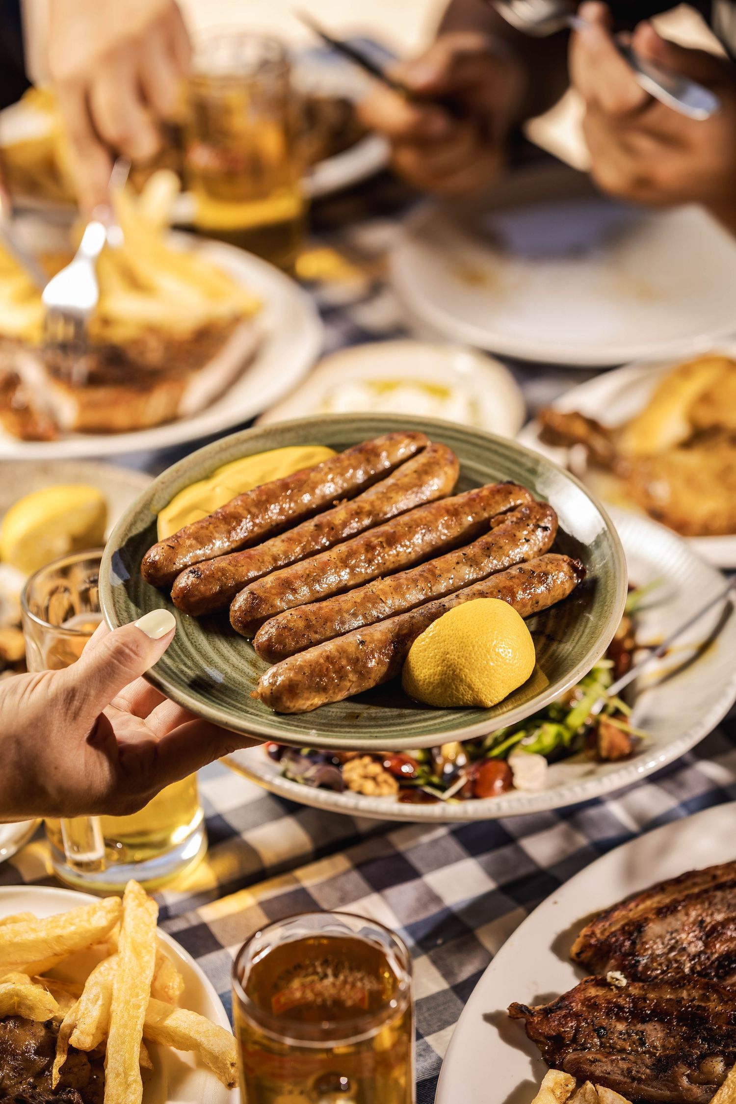 Grilled sausages served on plates with french fries and dipping sauce on a checkered tablecloth.