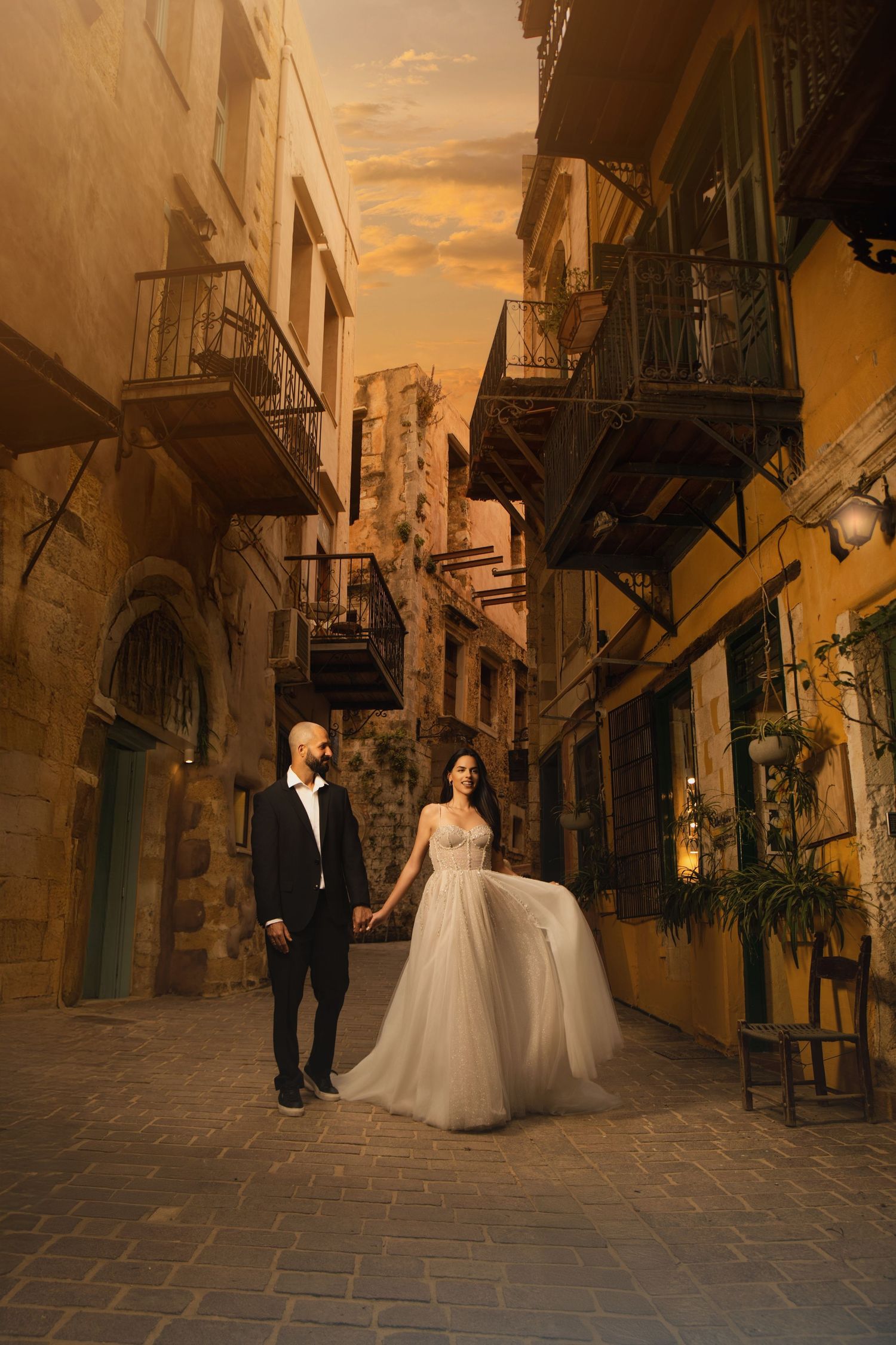 A couple in wedding attire strolling through charming European cobblestone streets at sunset.
