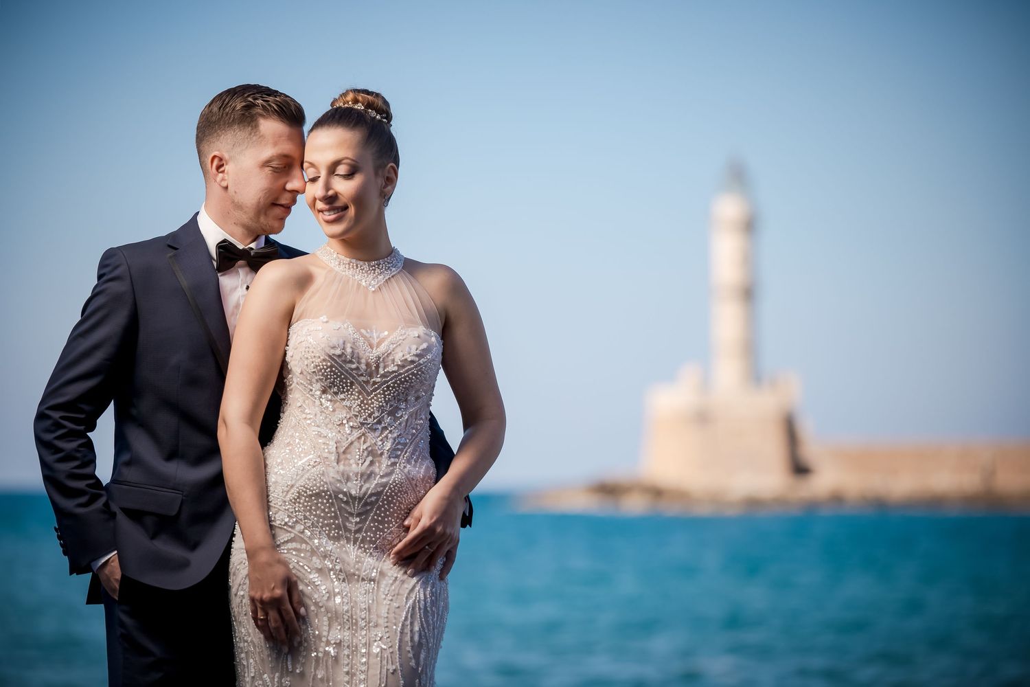 Elegant couple in wedding attire embracing near a seaside lighthouse under bright blue skies.