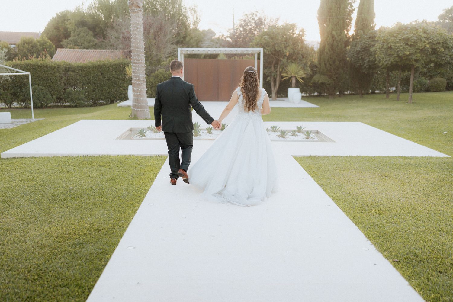 A wedding couple walks hand in hand down a white walkway in a sunny garden setting.