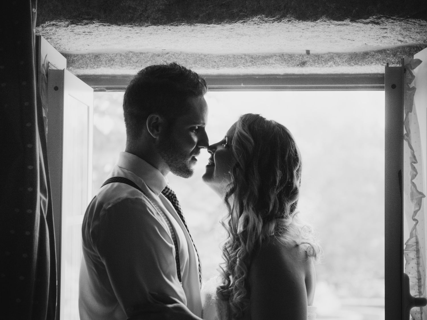 A romantic couple stands intimately close in silhouette by a window creating a tender black and white moment.
