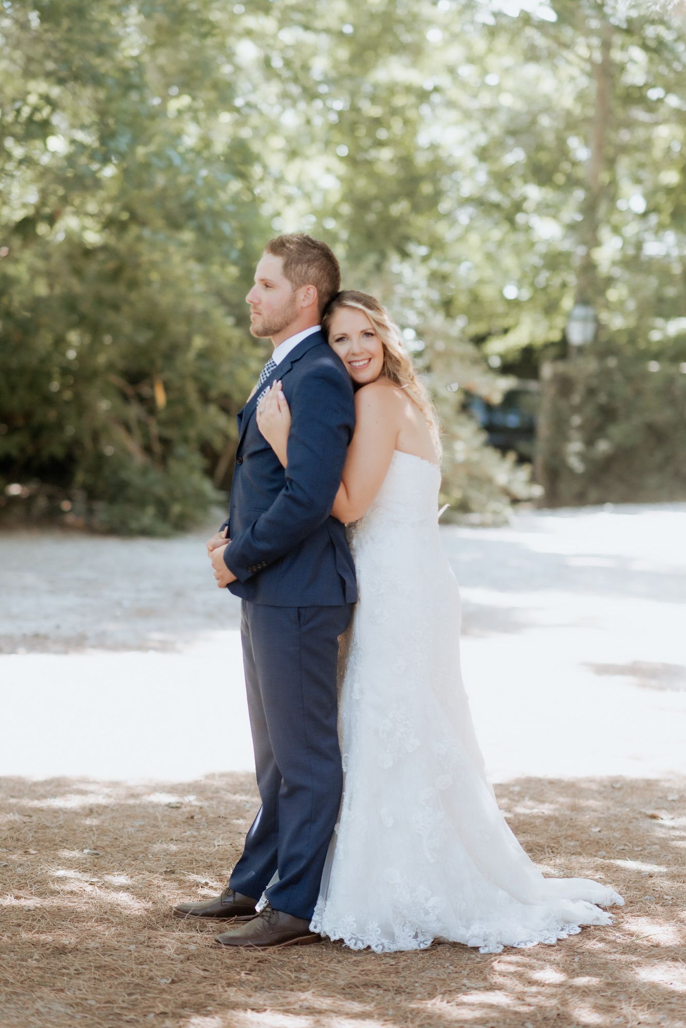 A couple in wedding attire stand back-to-back outdoors in a sun-dappled natural setting among trees.