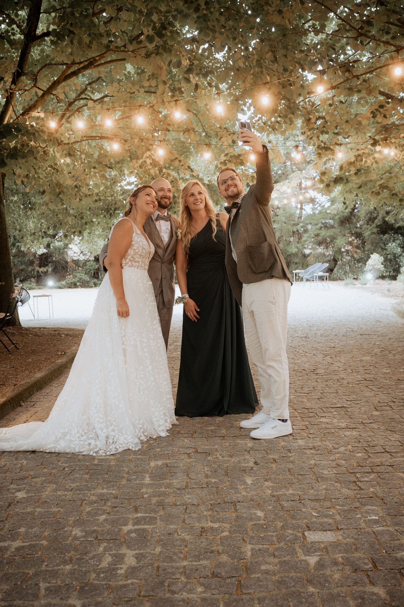 Wedding group celebration under string lights and tree canopy on a cobblestone path at dusk.
