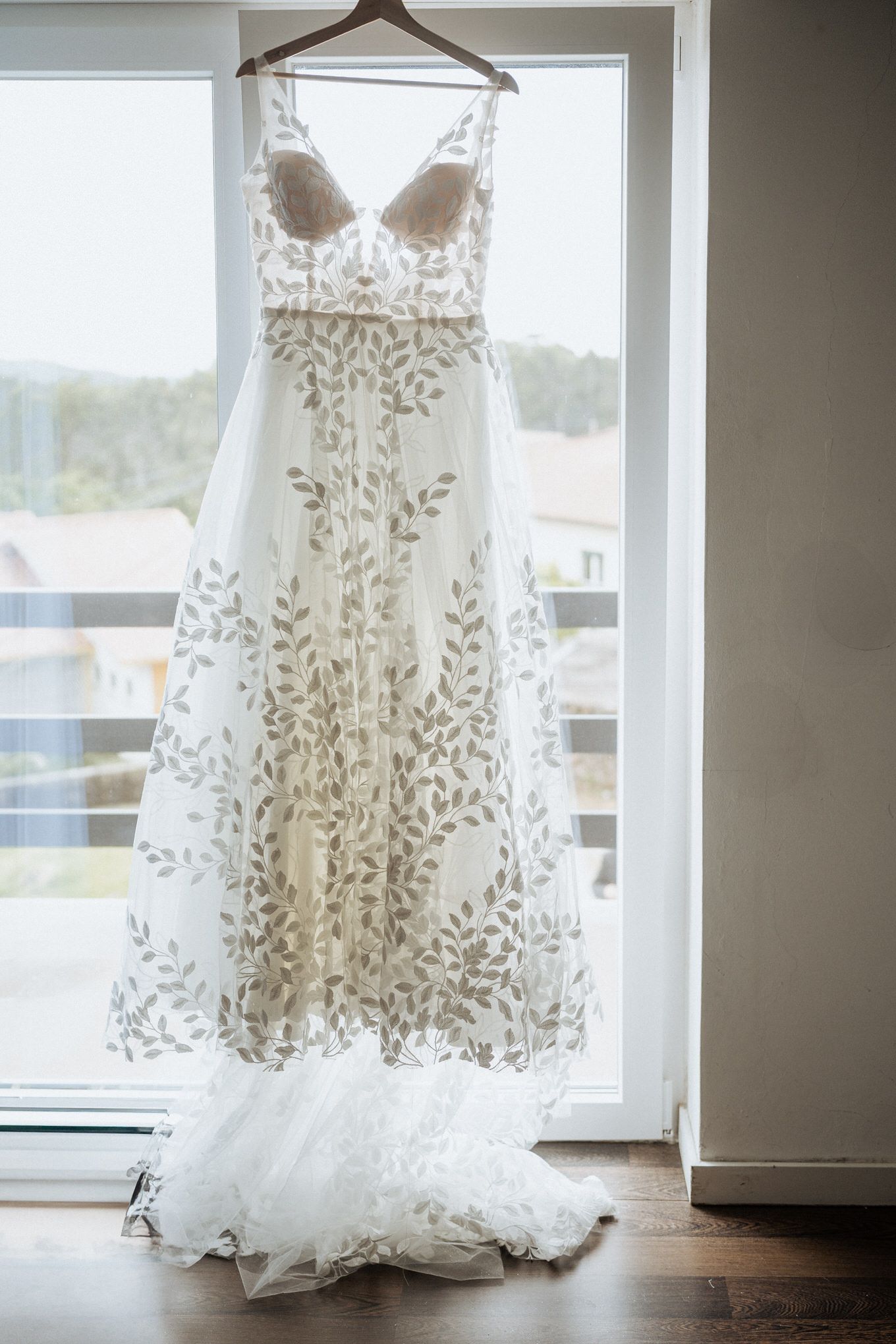 Elegant white wedding dress with delicate floral lace embroidery hangs by a window, showcasing its flowing silhouette and train.