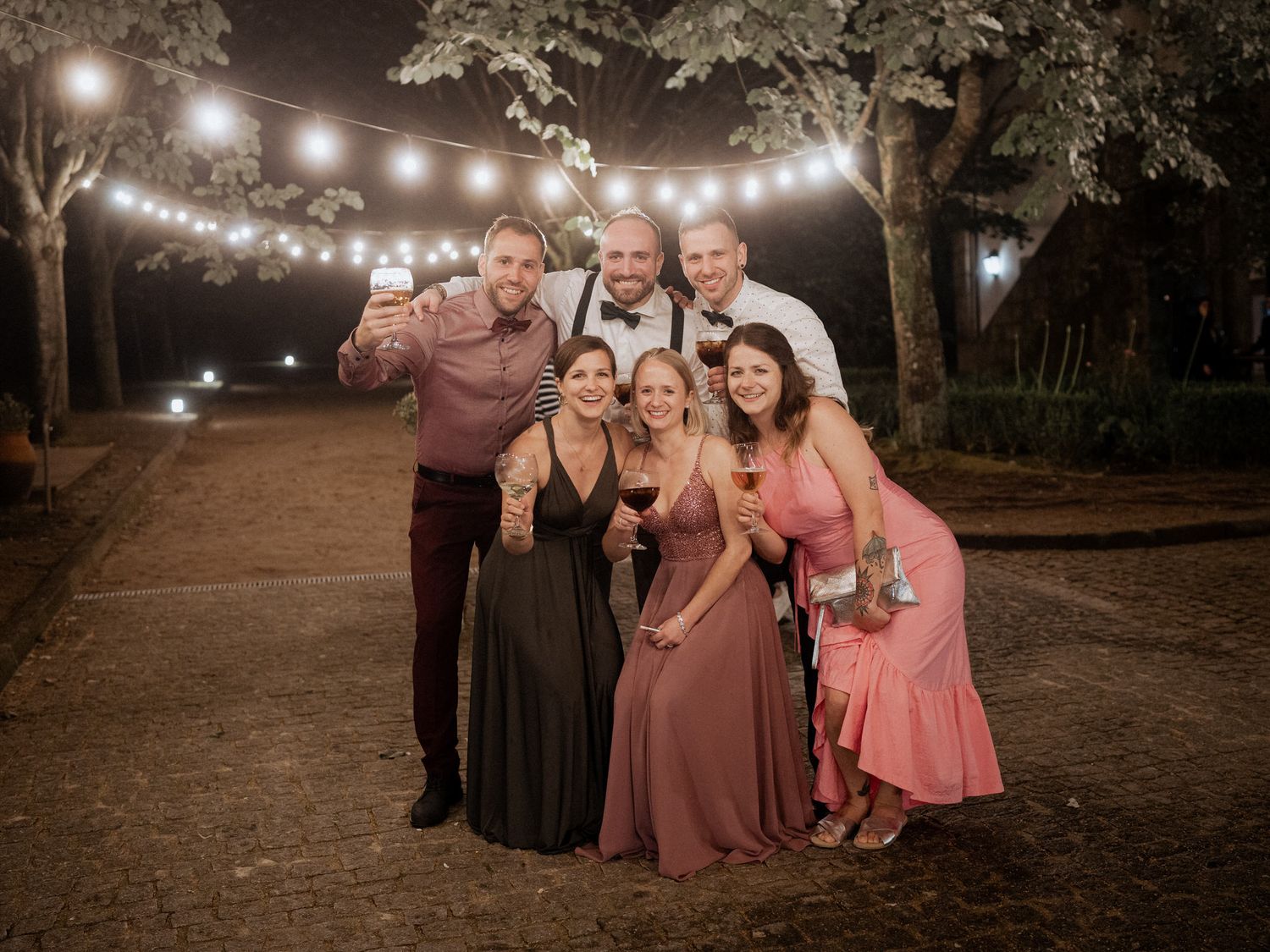 A group poses together under warm string lights on a dirt path during a special evening celebration.