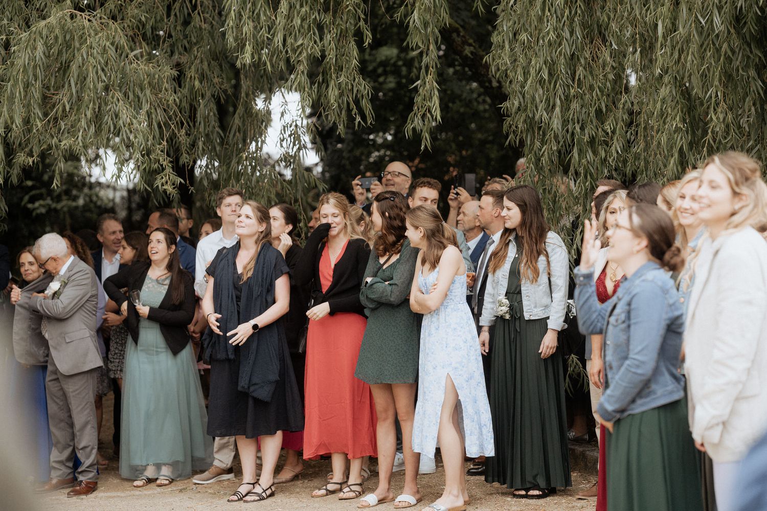 Group of guests in formal attire gather outdoors under a weeping willow tree during a wedding ceremony.