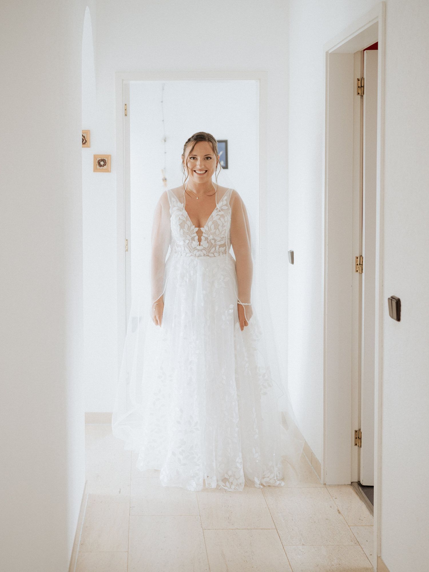 Beautiful white lace wedding gown with flowing skirt and illusion sleeves photographed in bright interior hallway.