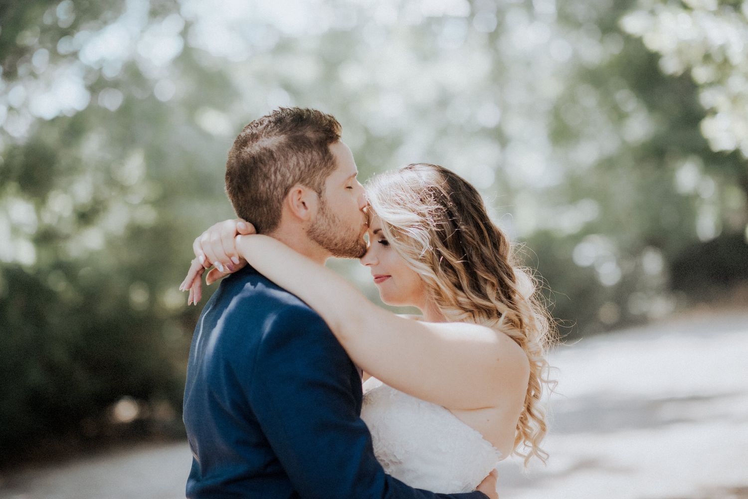 A couple in wedding attire share a romantic kiss outdoors with a soft bokeh background.