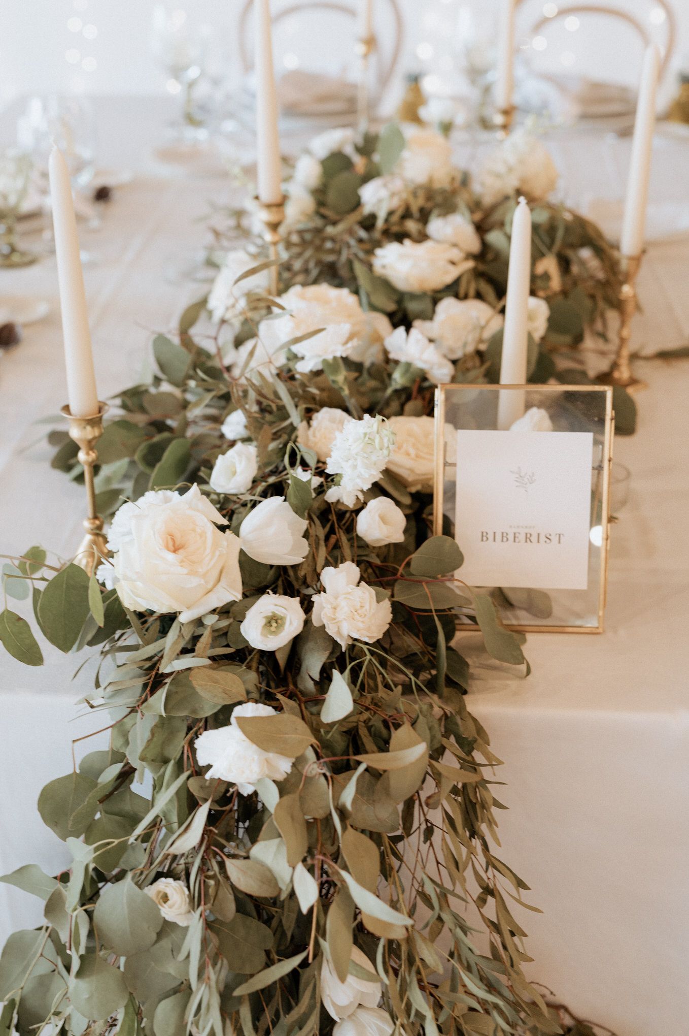 Elegant table decor featuring white roses and eucalyptus garland with tall taper candles in gold holders.