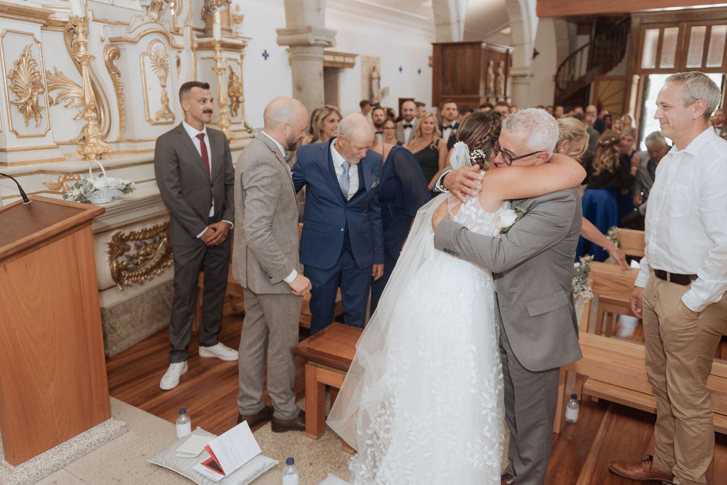 Wedding ceremony taking place inside an ornate church with guests gathered around the couple during an emotional embrace.