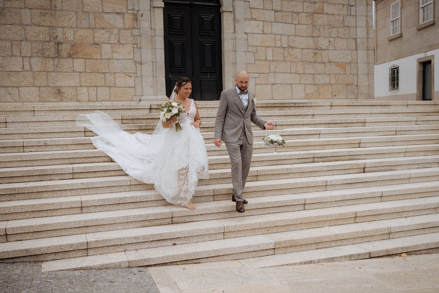 A joyful bride in a flowing white dress descends stone stairs with her groom in a gray suit.