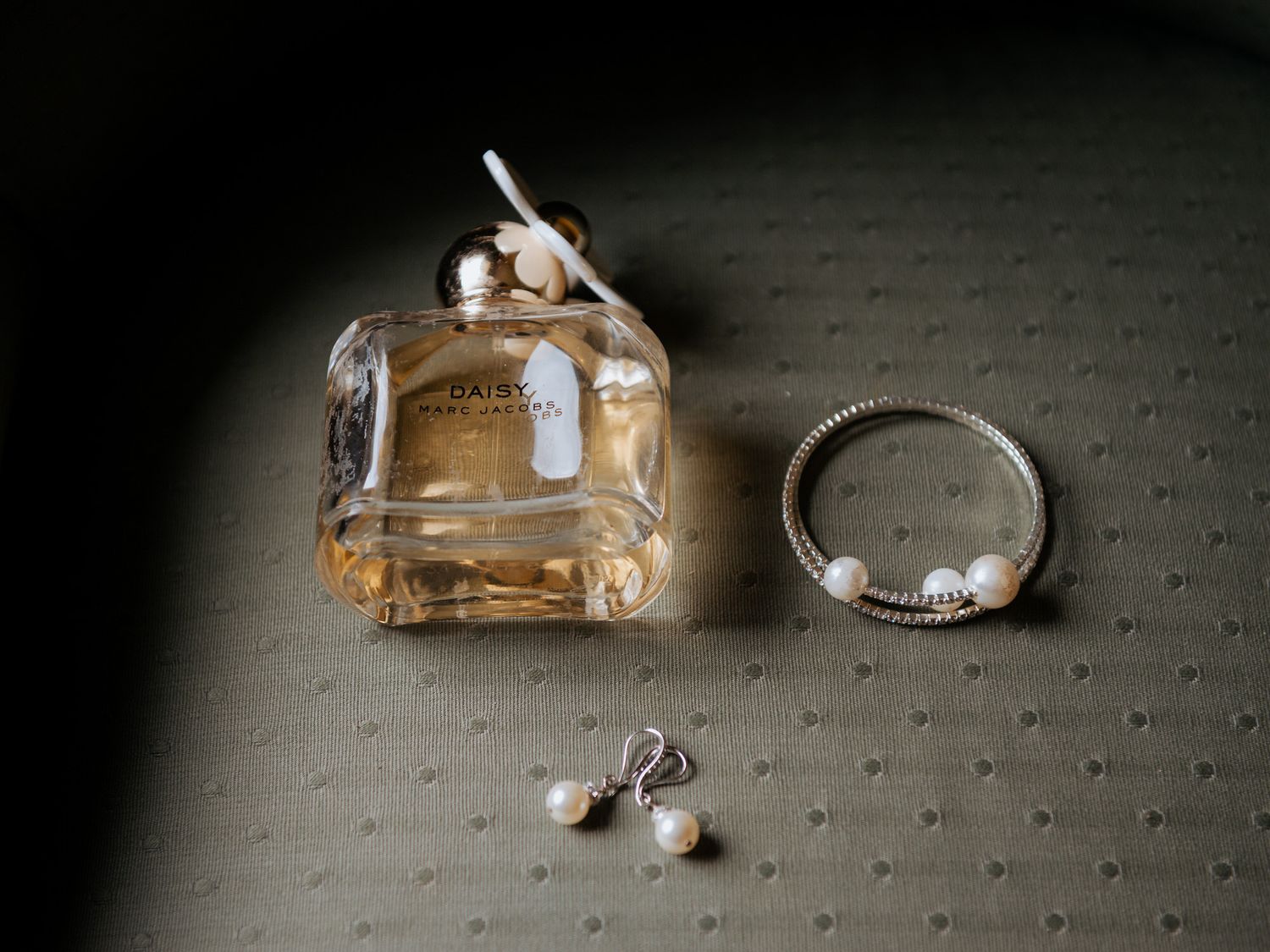 Wedding perfume bottle, delicate diamond ring, and pearl earrings arranged on a textured gray surface.
