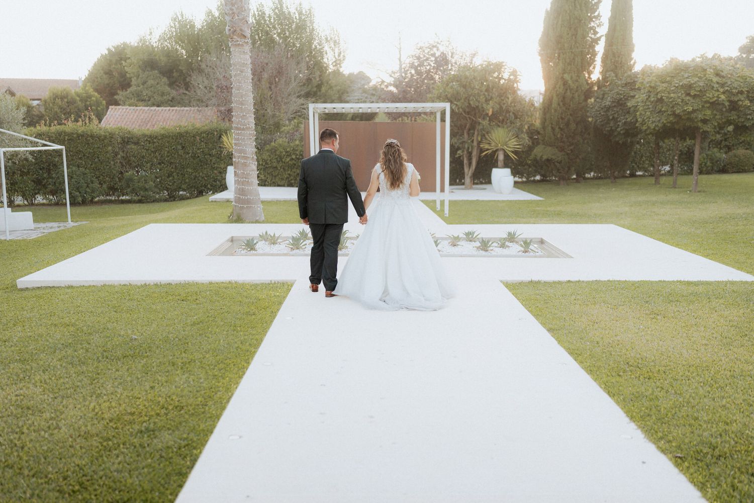 Couple walks down white outdoor aisle on grassy lawn with vaulted ceremony space in background.