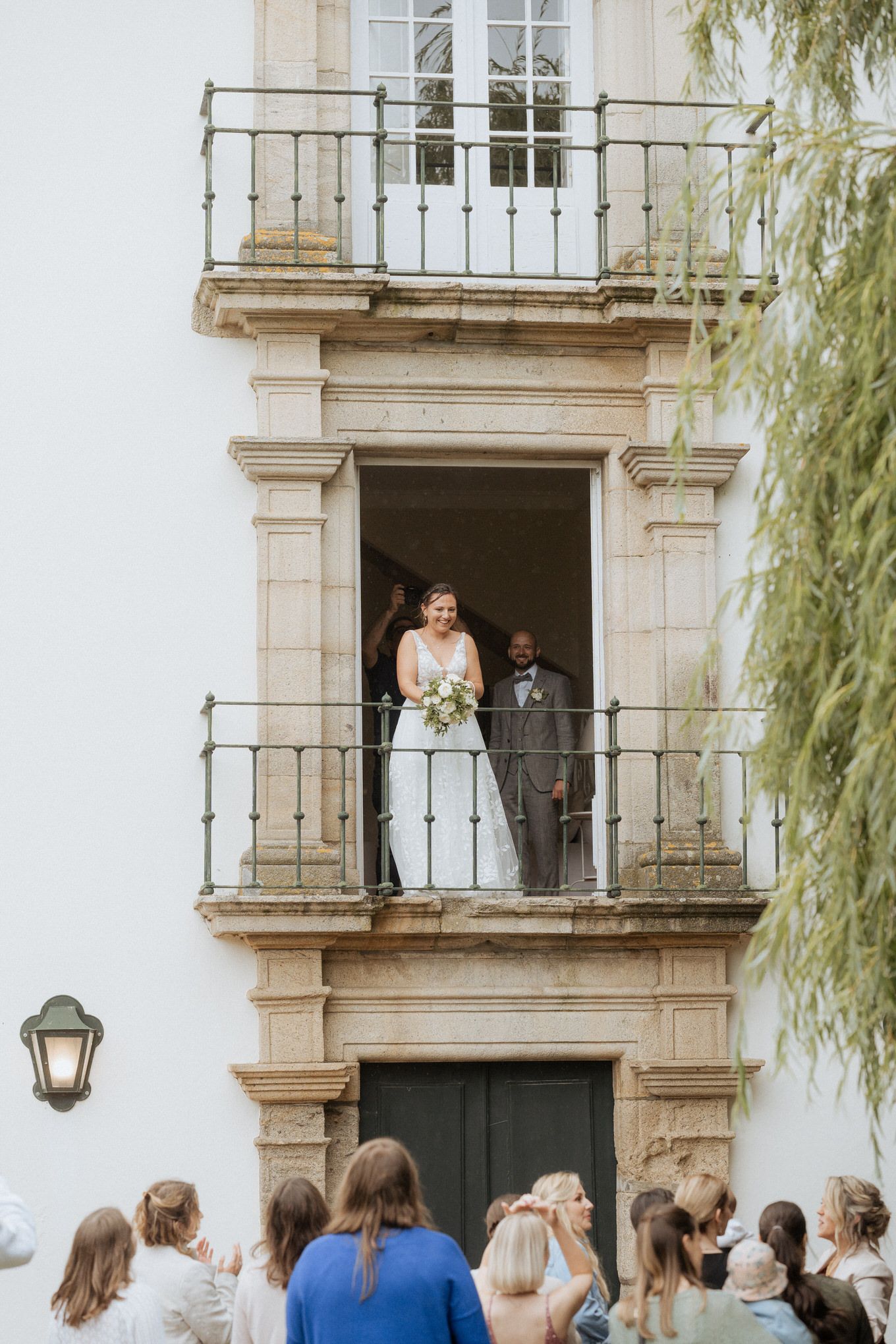 Wedding guests gather below a historic balcony with white flowers and willow trees in view.