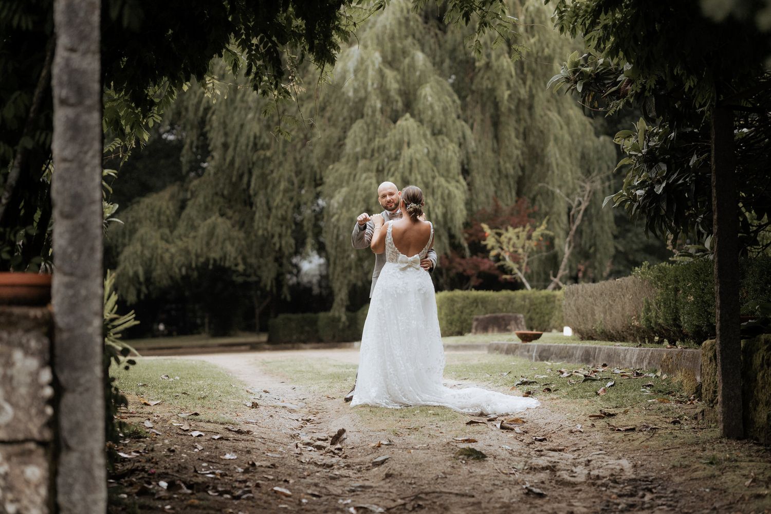 A person in a white wedding dress stands on a garden path surrounded by willow trees on a cloudy day.