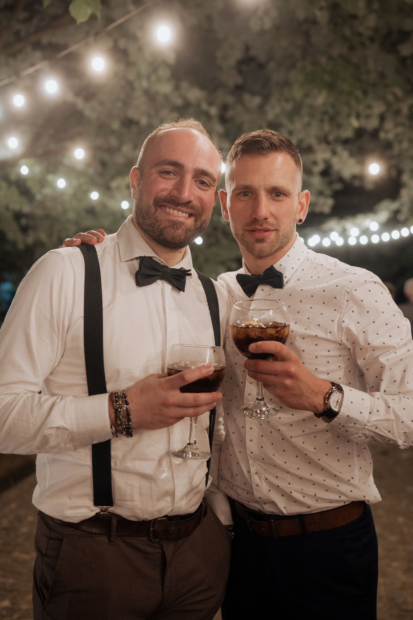 Two formally dressed wedding guests smile and toast with cocktail drinks under outdoor string lights at evening celebration.