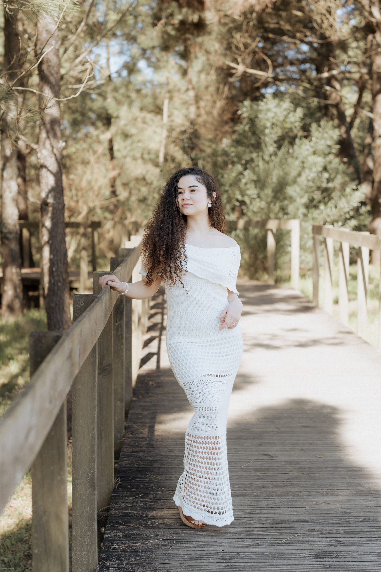 Mulher com vestido branco de croché esvoaçante posa num caminho de tábuas de madeira através de uma zona arborizada sob uma luz suave do sol
