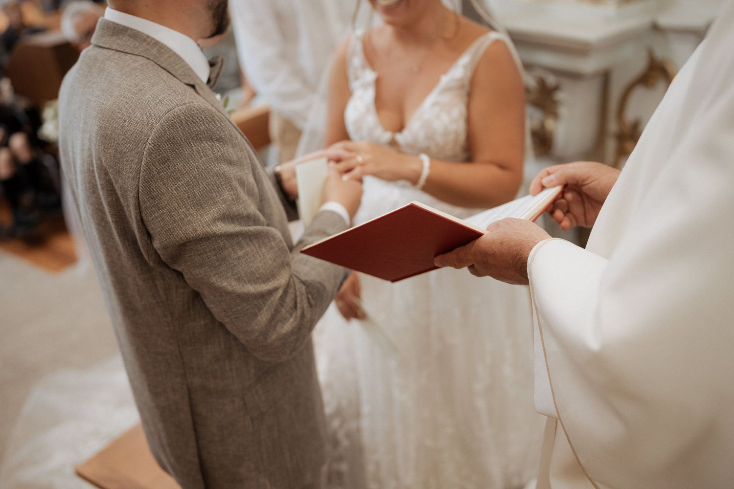 A wedding ceremony shows hands holding a burgundy book during the exchange of vows in an elegant indoor setting.