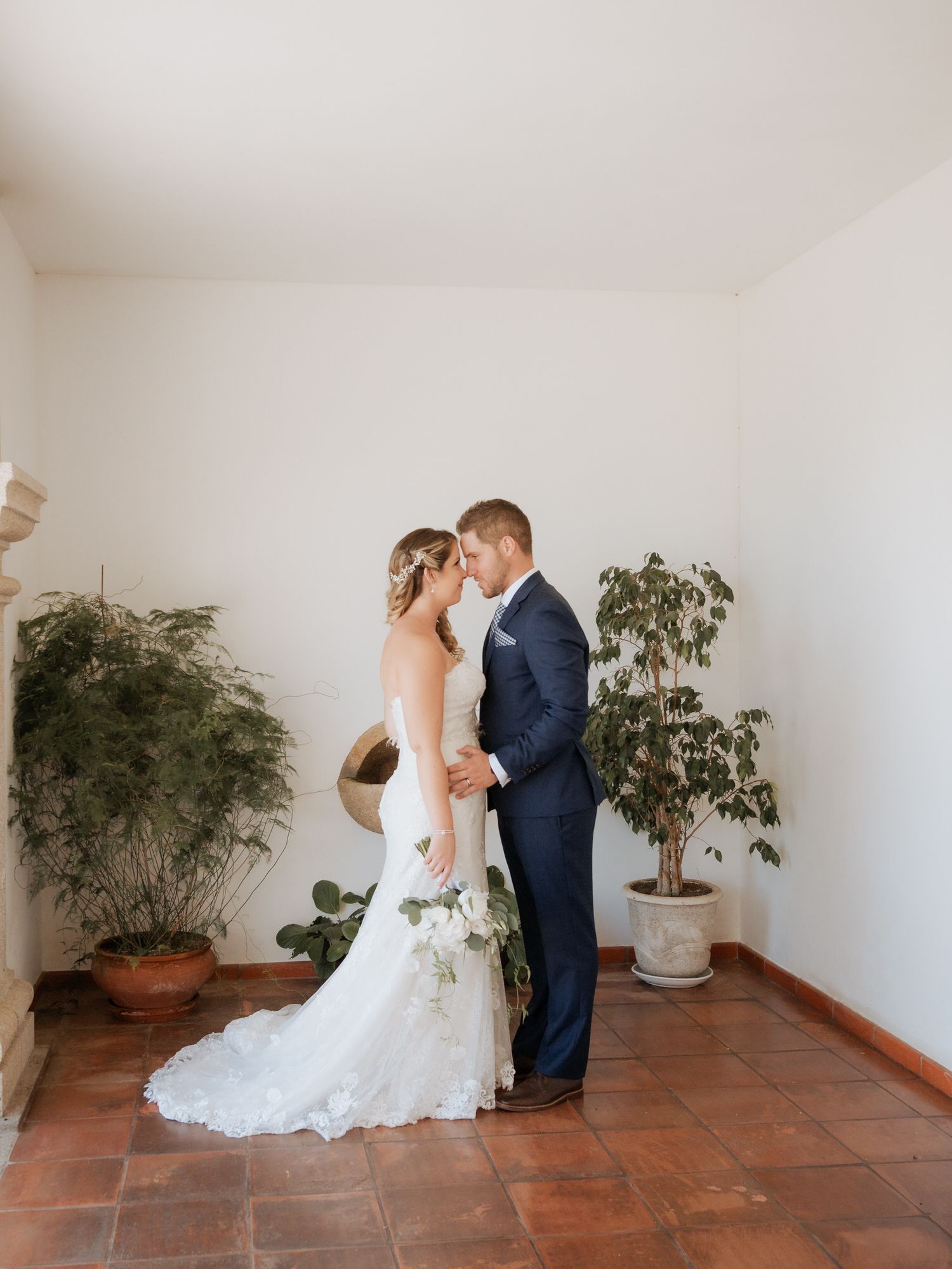 Romantic couple stands together on terracotta tile floor with potted plants in an elegant indoor setting.