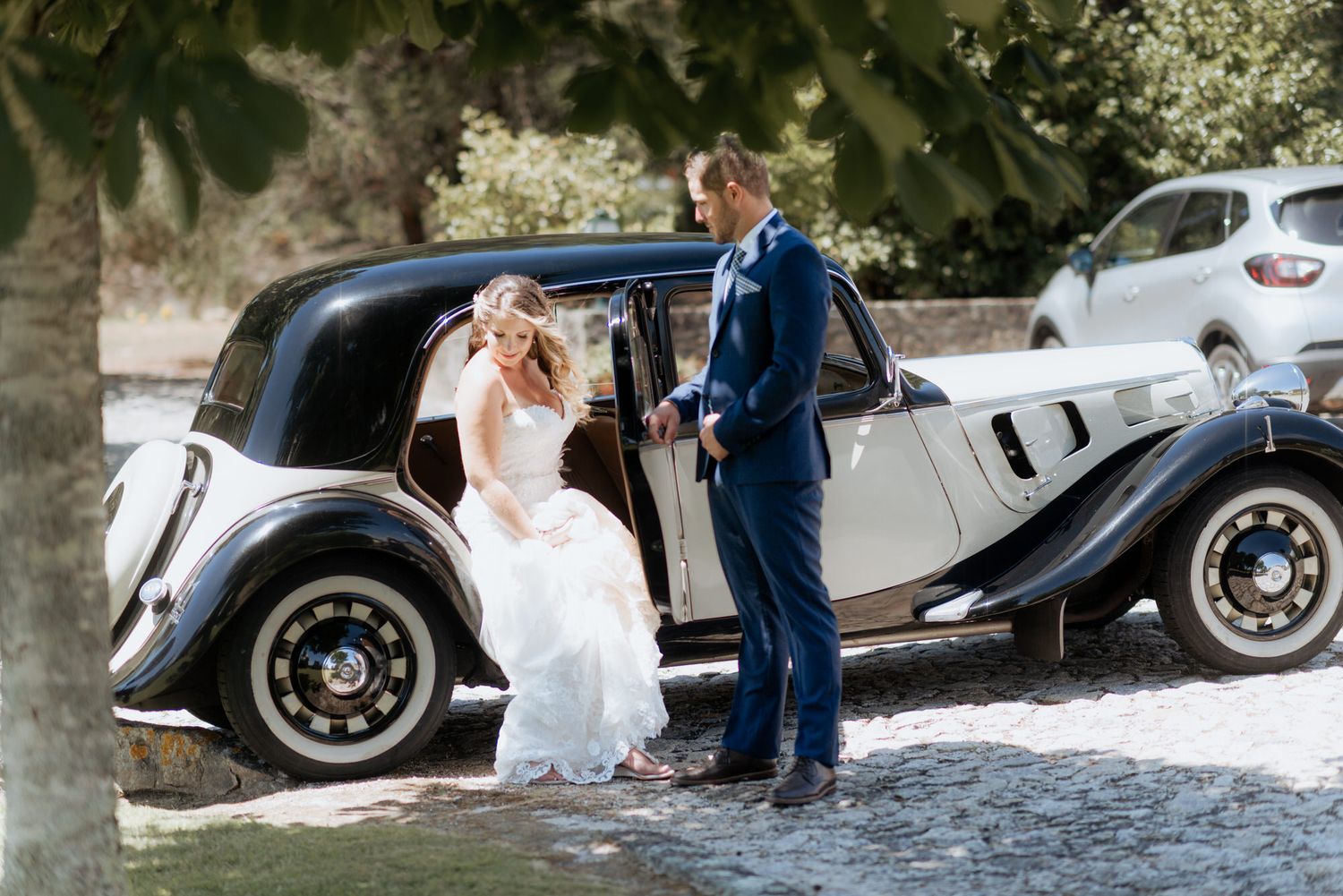 A couple stands next to a vintage grey car in an elegant wedding setting under trees.