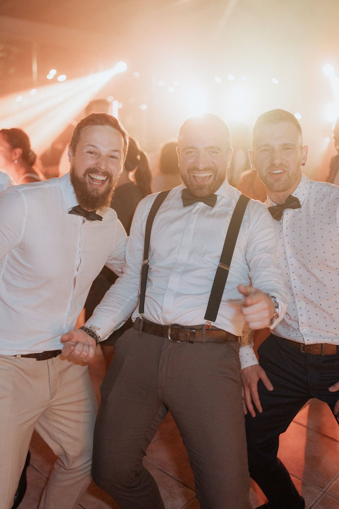 Three men in formal attire with bow ties and suspenders smile together at a festive party with bright lights in background.