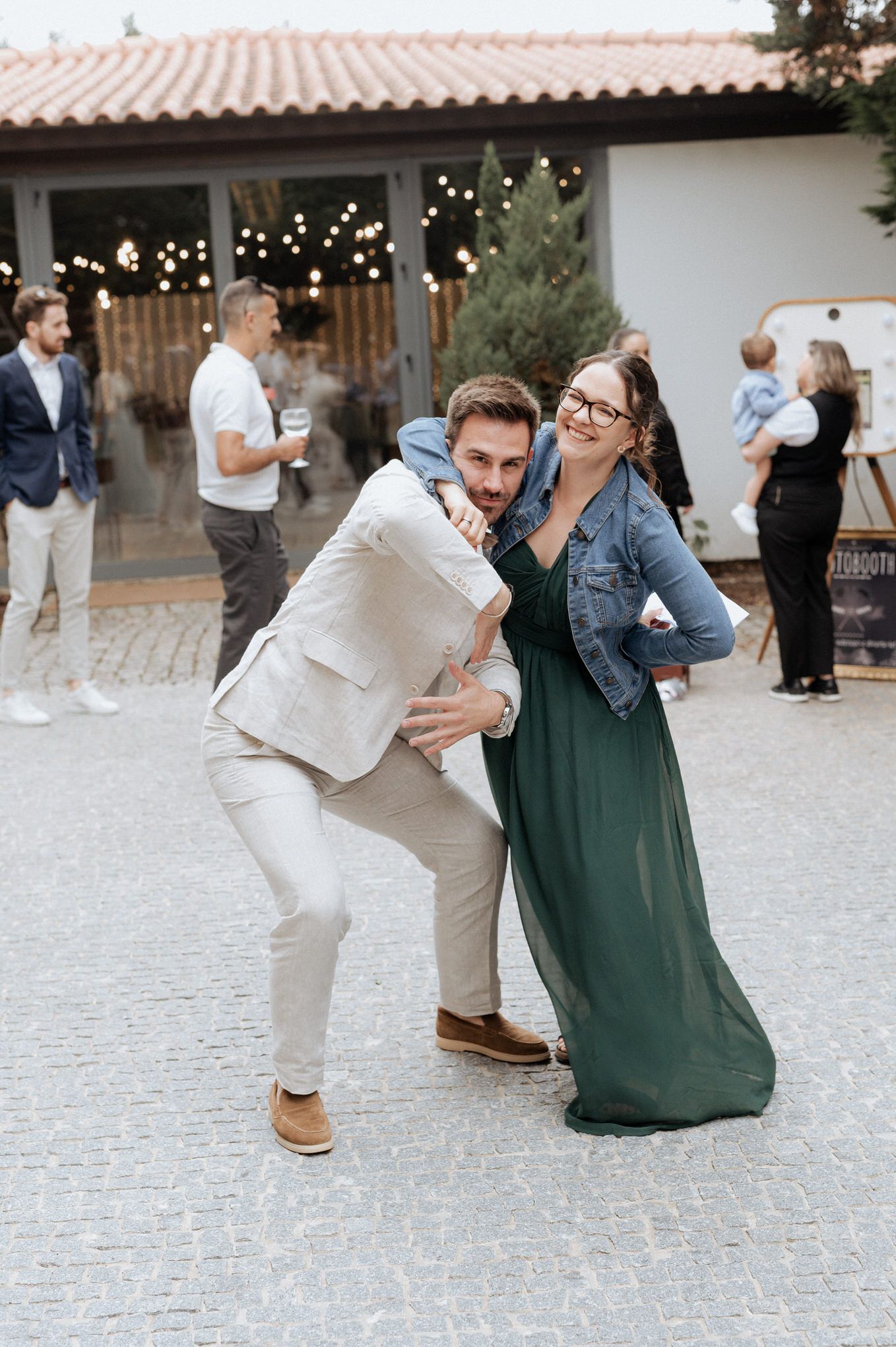 A couple striking a playful pose at an outdoor party venue with string lights and guests in the background.