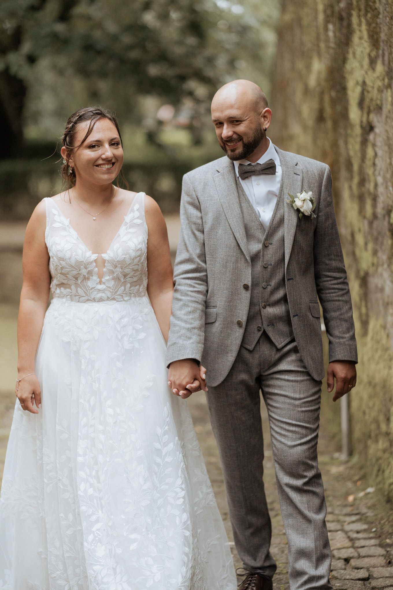 A couple in elegant wedding attire walking hand in hand along a cobblestone path in a garden setting.