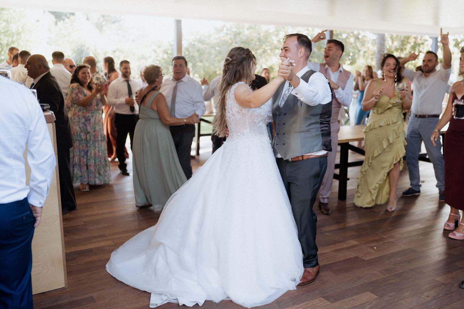 Newlyweds and wedding guests dance together on a wooden floor under a covered outdoor venue during reception celebrations.