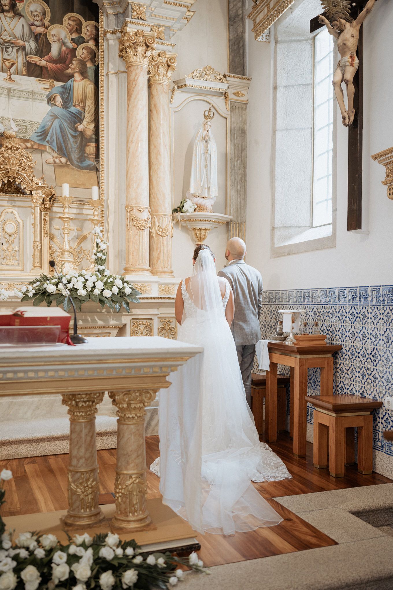 A couple stands reverently at the altar of an ornate Catholic church decorated with white floral arrangements and religious statues.