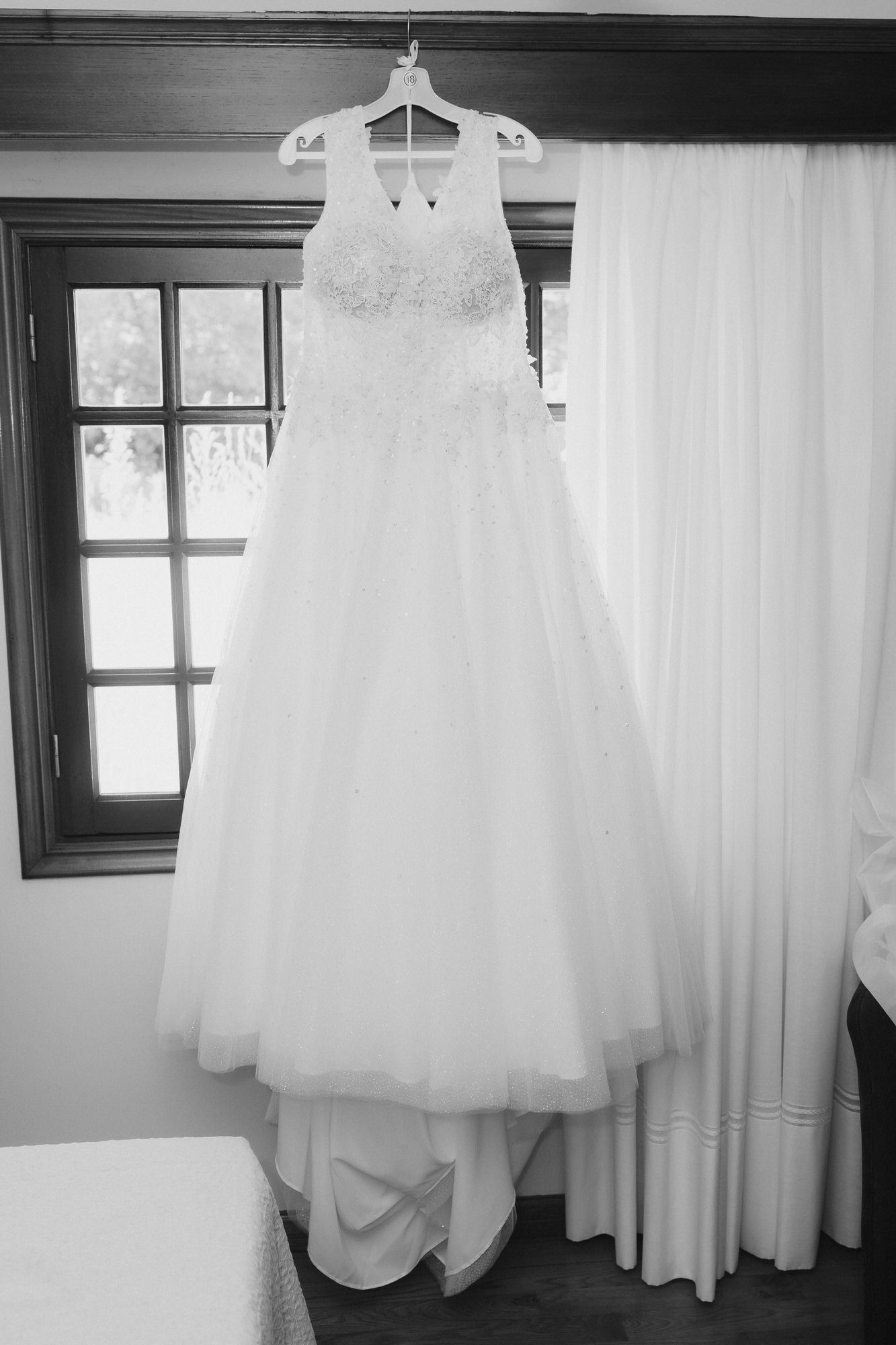 A classic white lace wedding dress with full tulle skirt hangs on display near a window.