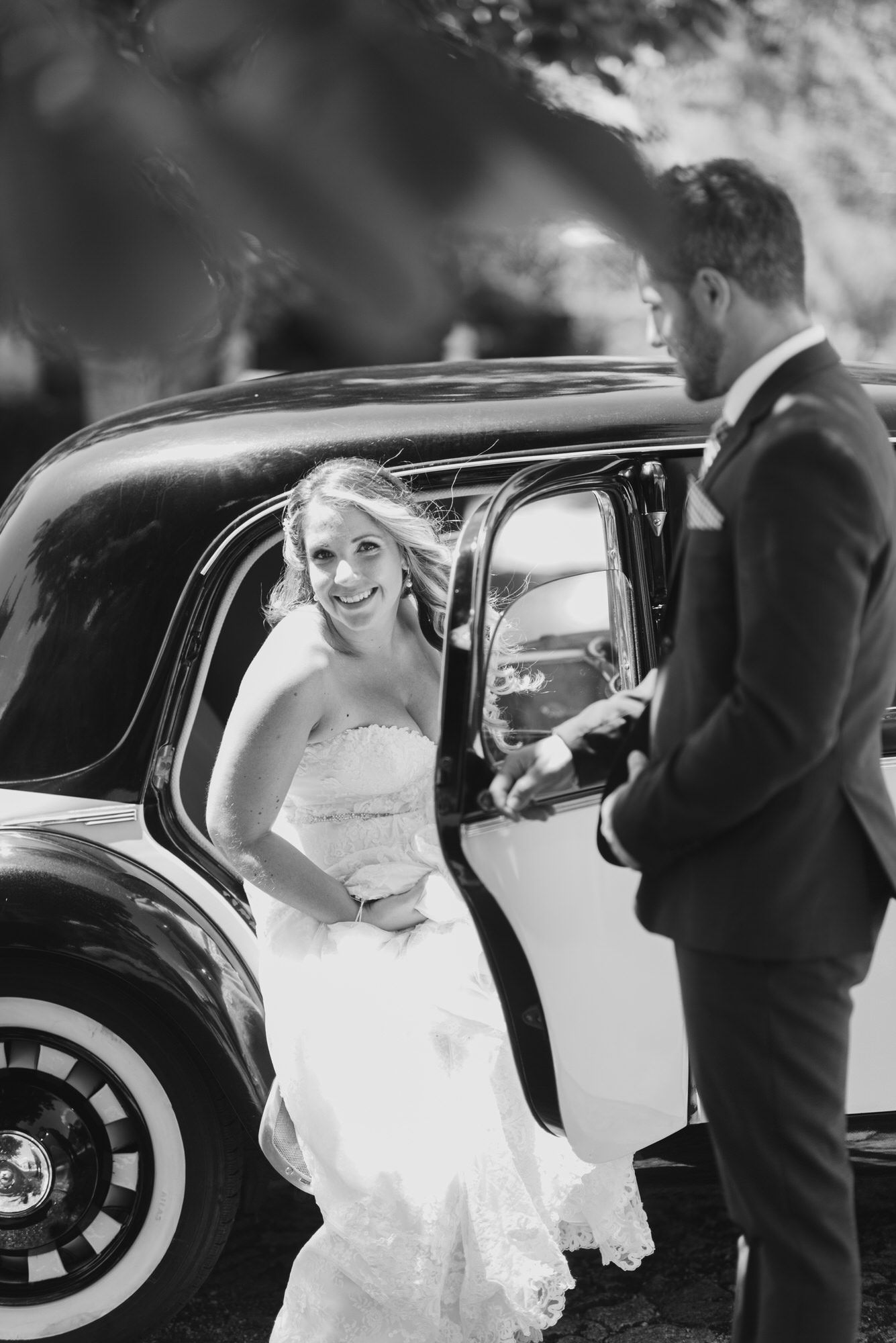 Black and white wedding photo showing a couple with a vintage car, with the bride in a white dress getting into the vehicle.